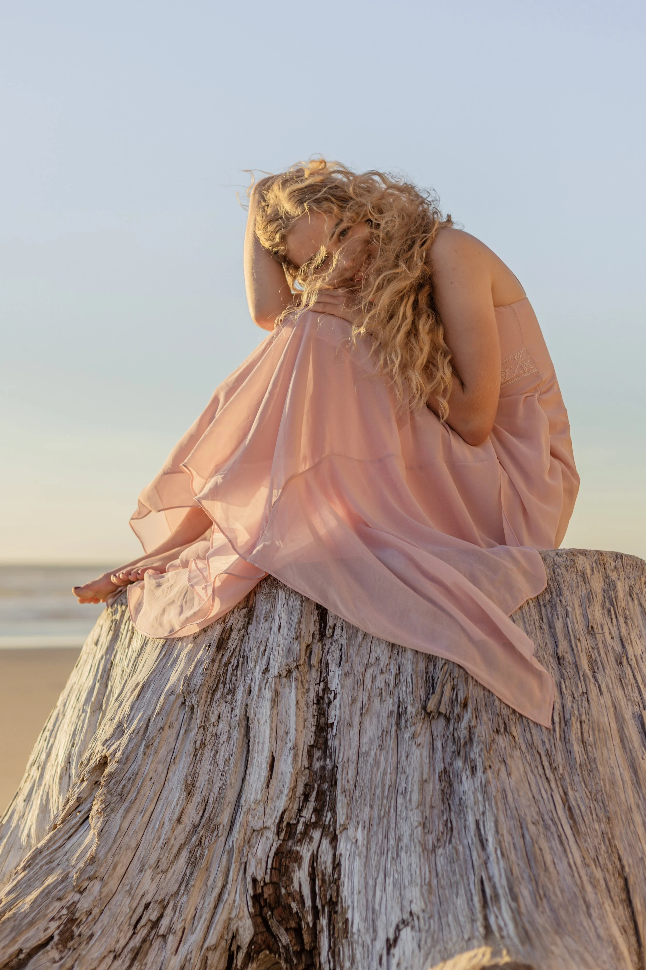 A woman with long curly blonde hair sitting on a large weathered piece of driftwood on a beach at sunset, wearing a flowing pink dress.