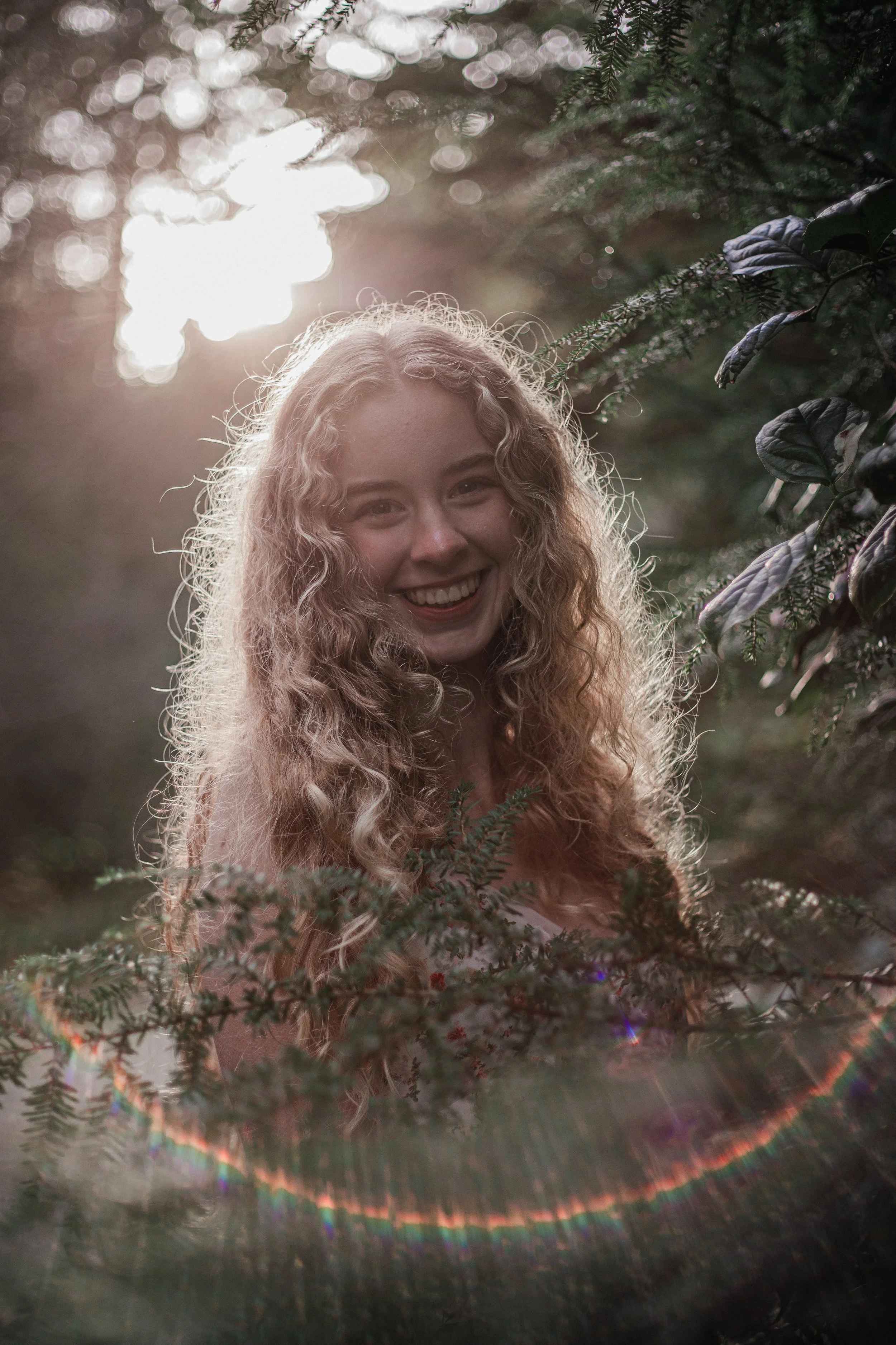A woman with long, curly blonde hair smiling outdoors among green foliage with sunlight in the background.