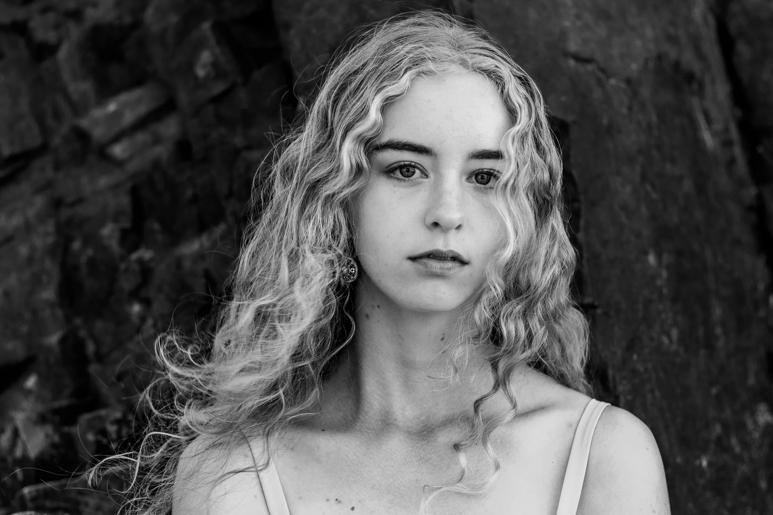 Black and white portrait of a young woman with curly hair, wearing earrings and a spaghetti strap top, standing in front of a rocky background.