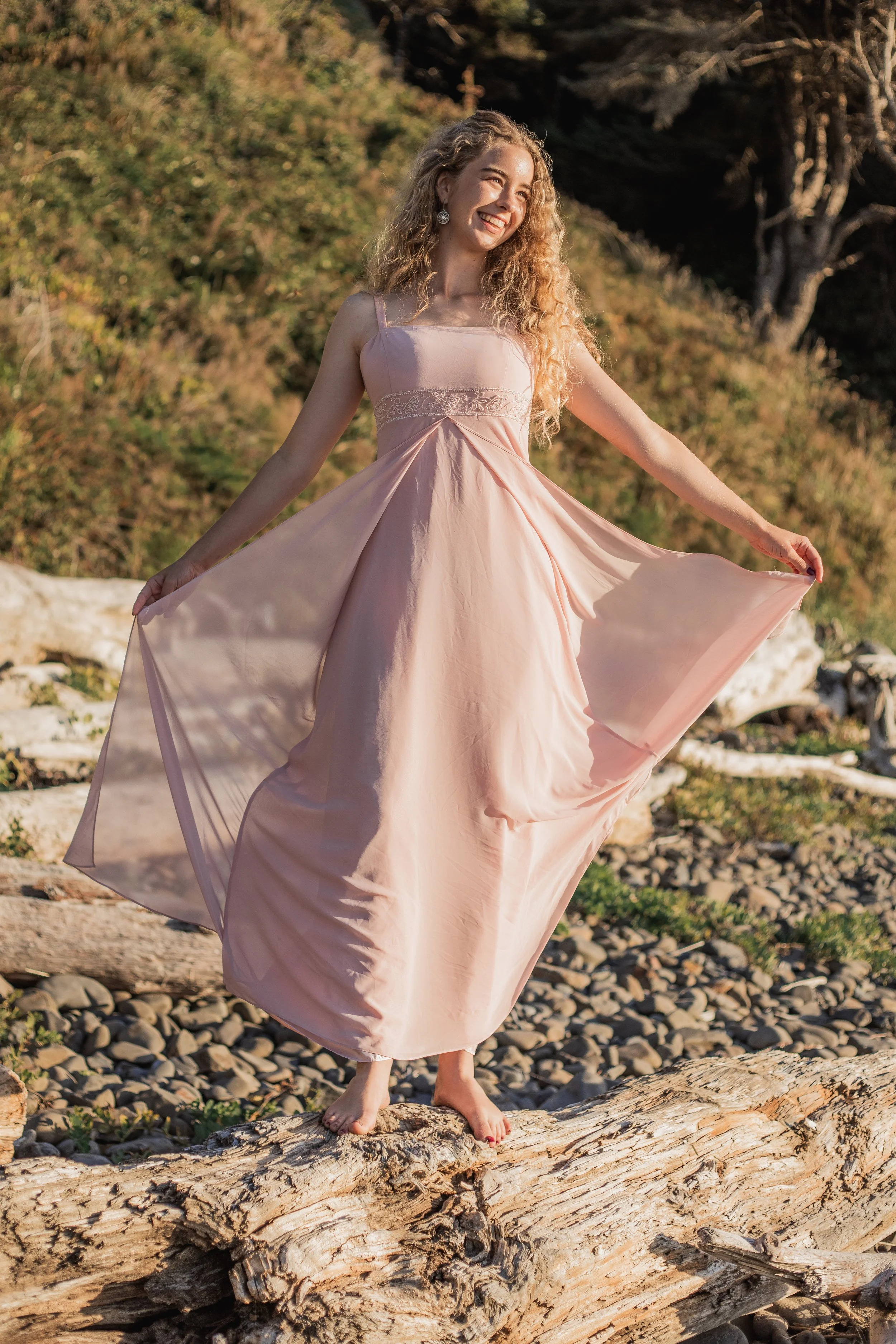 A young woman with curly blonde hair in a flowing pink dress standing barefoot on a large driftwood log outdoors.