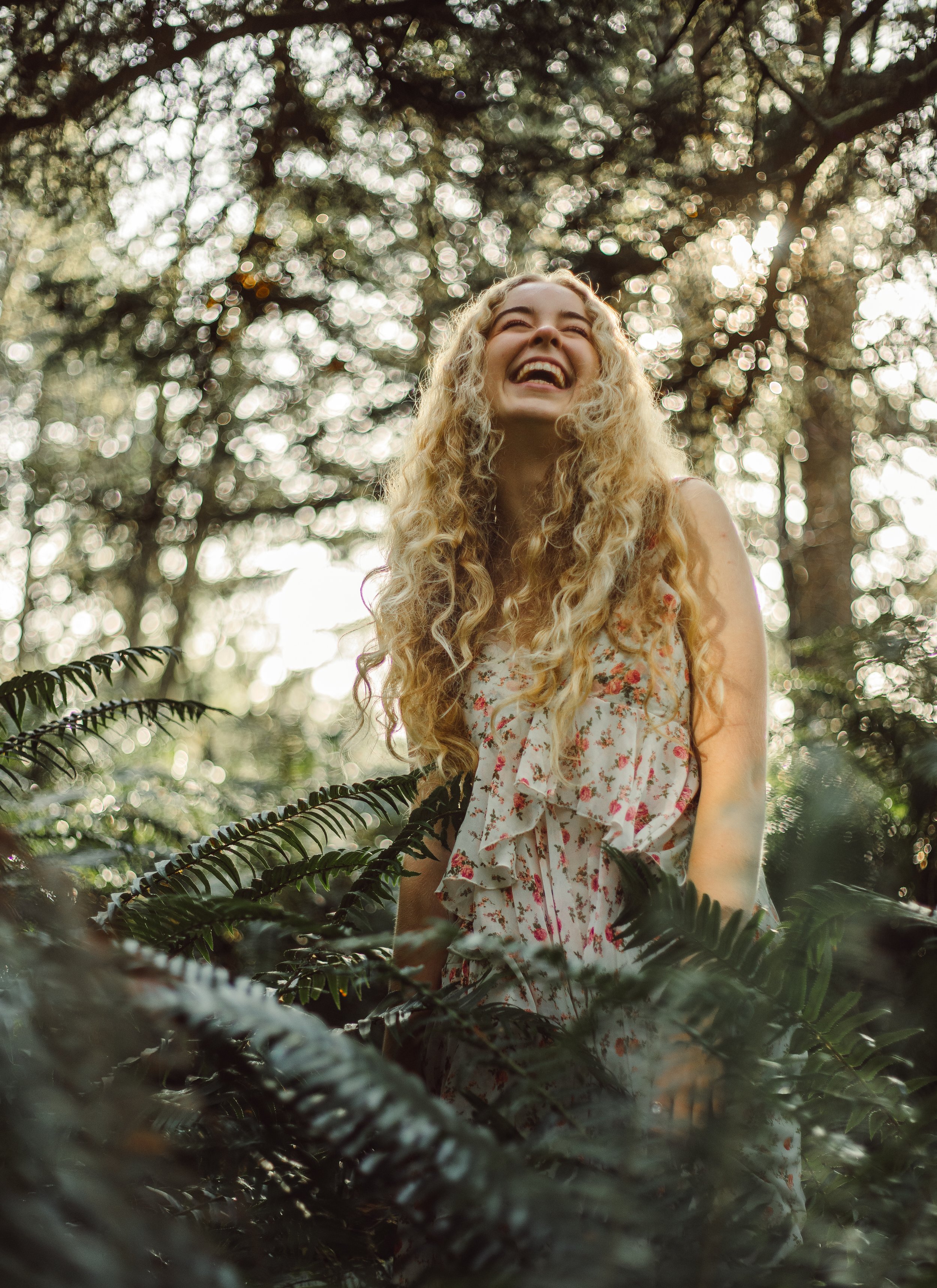 A young woman with long, curly blonde hair standing among lush ferns in a sunlit forest, smiling and looking joyful.