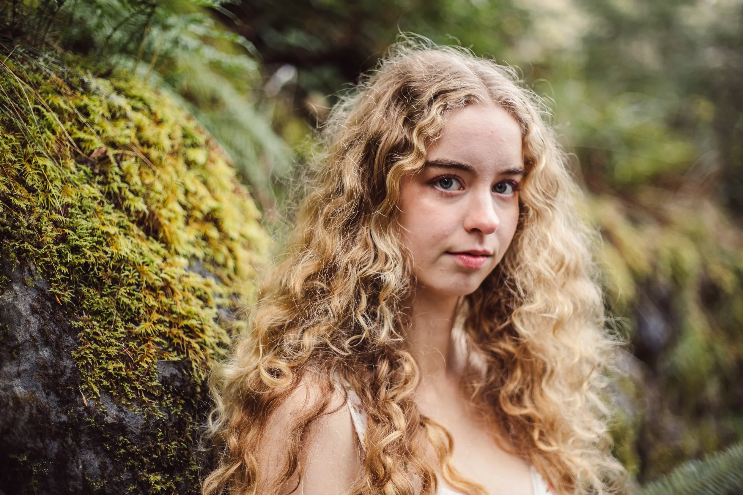 Young woman with long, curly blonde hair outdoors in a lush, green forest.