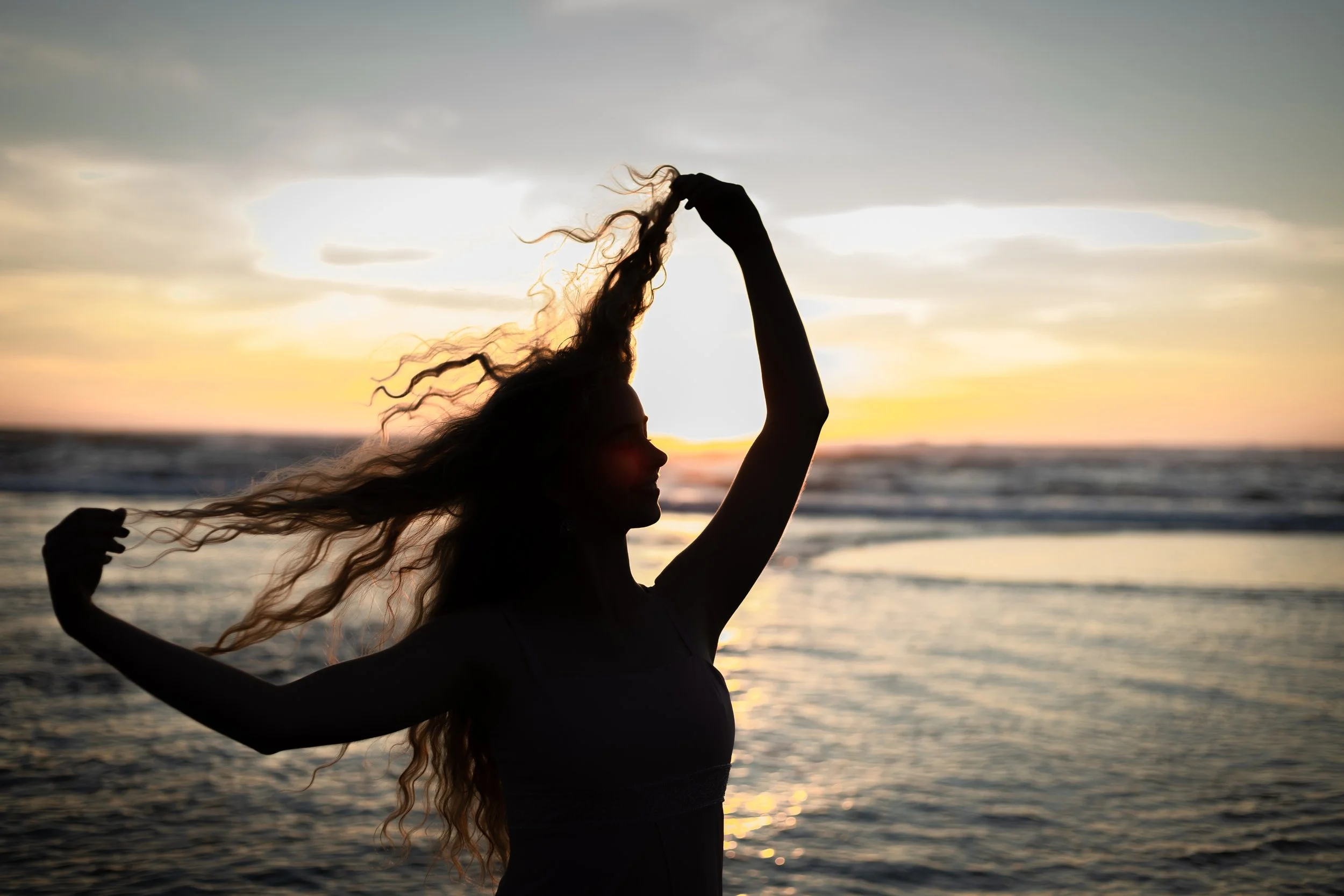 Silhouette of a woman with long, curly hair playing with her hair on a beach during sunset.