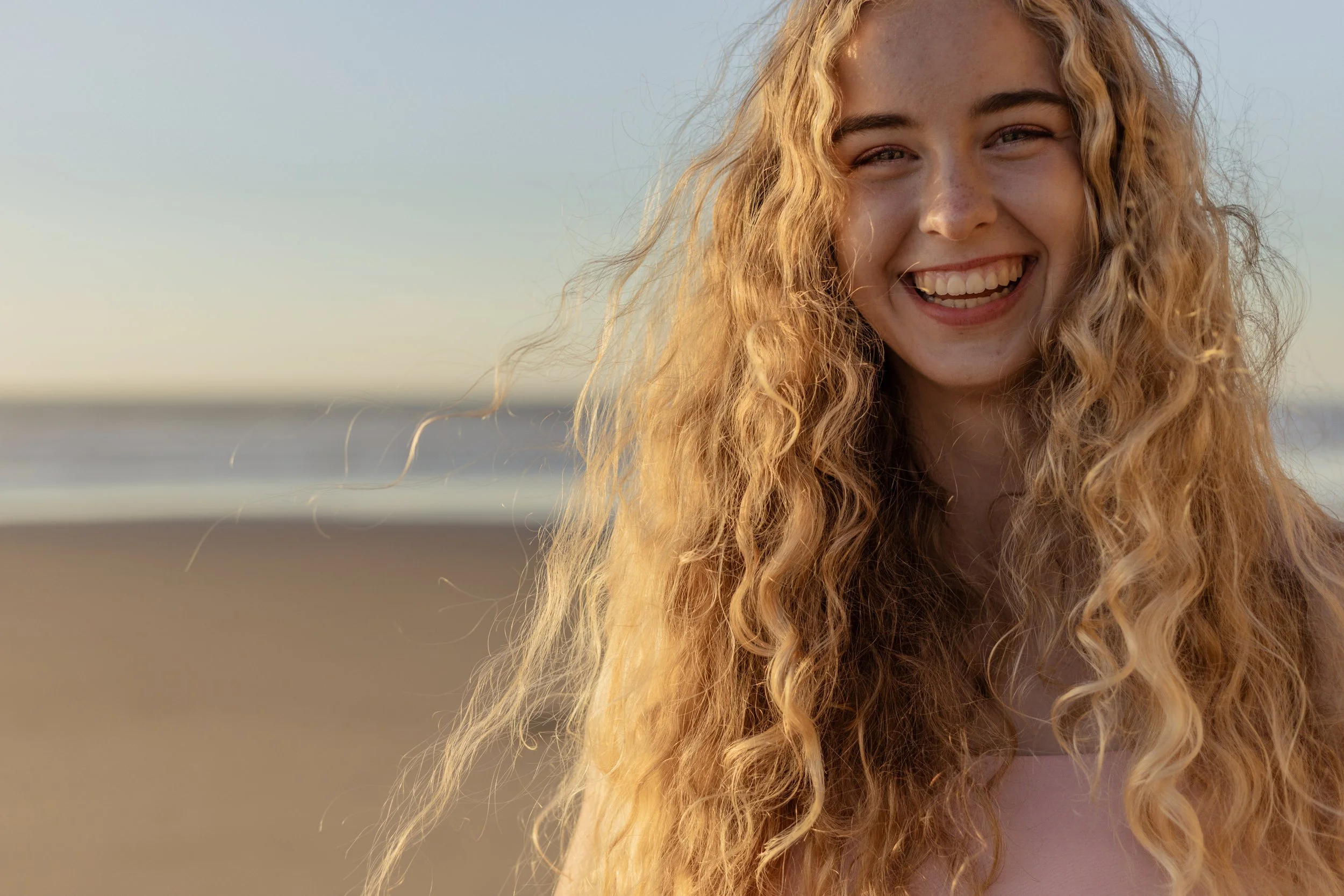 A young woman with long, curly blonde hair smiling on the beach during sunset.