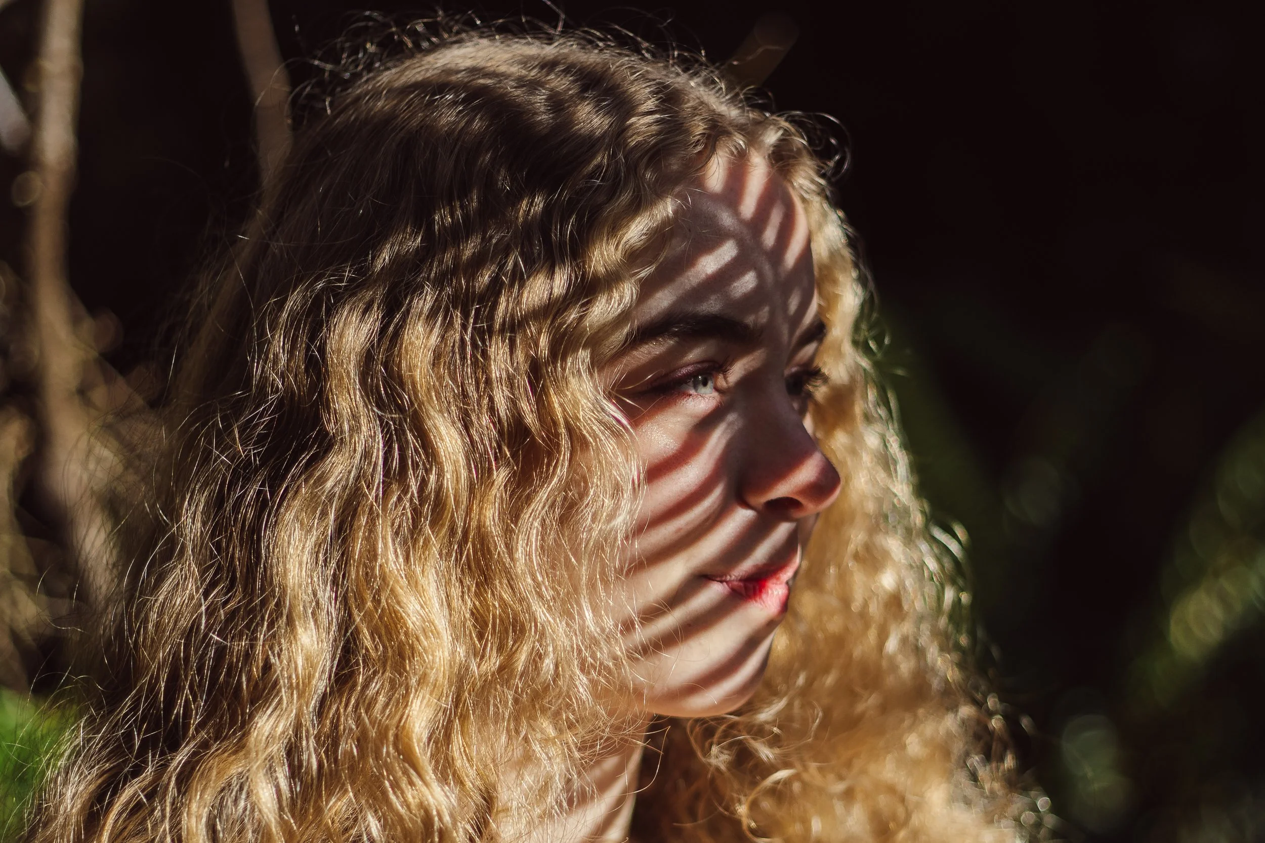 A woman with wavy blonde hair looking to the right, with shadows from a striped pattern cast on her face and hair in sunlight.