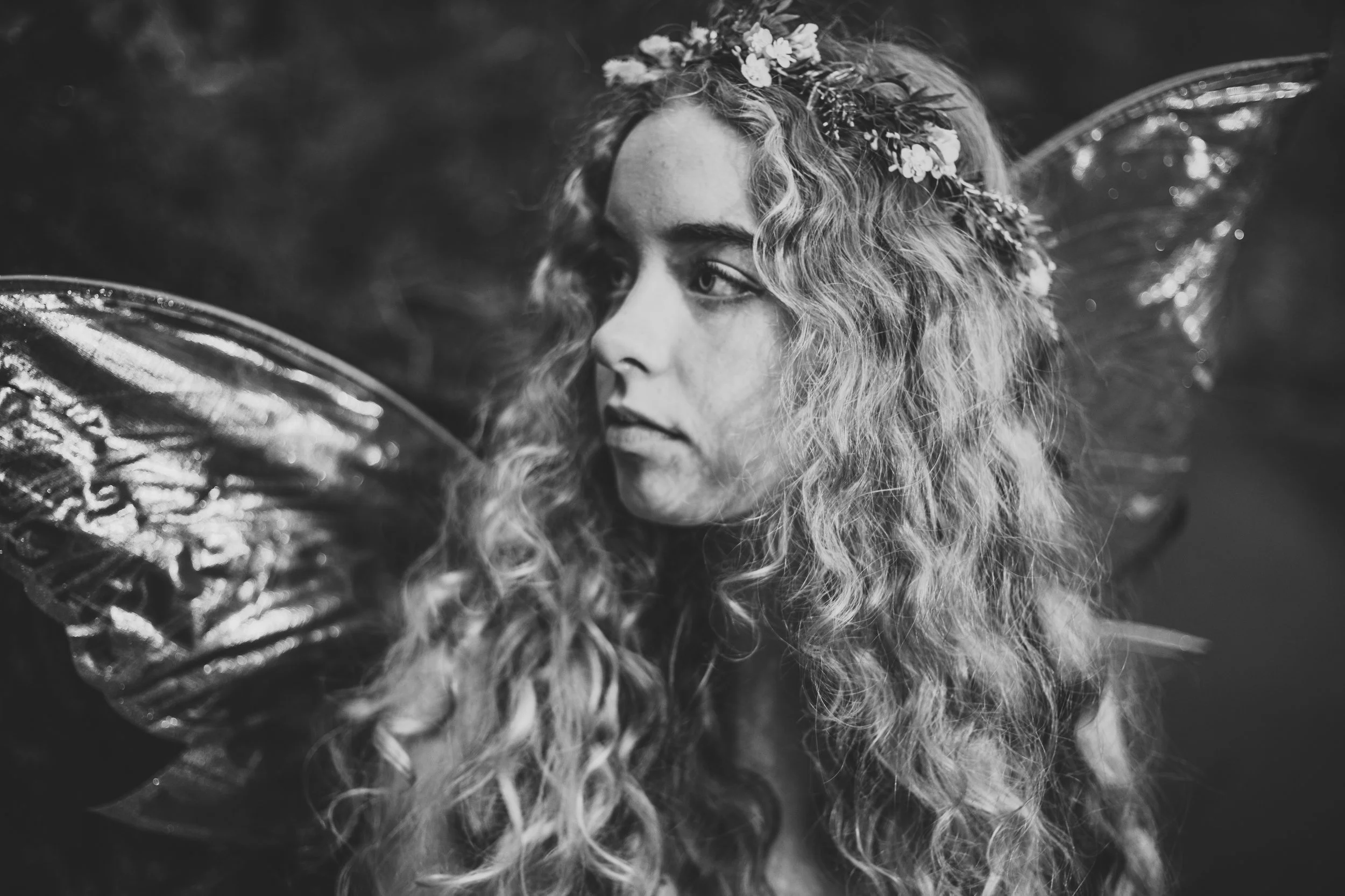 A woman with long curly hair wearing a floral crown and fairy wings, looking to the side in a black and white photo.