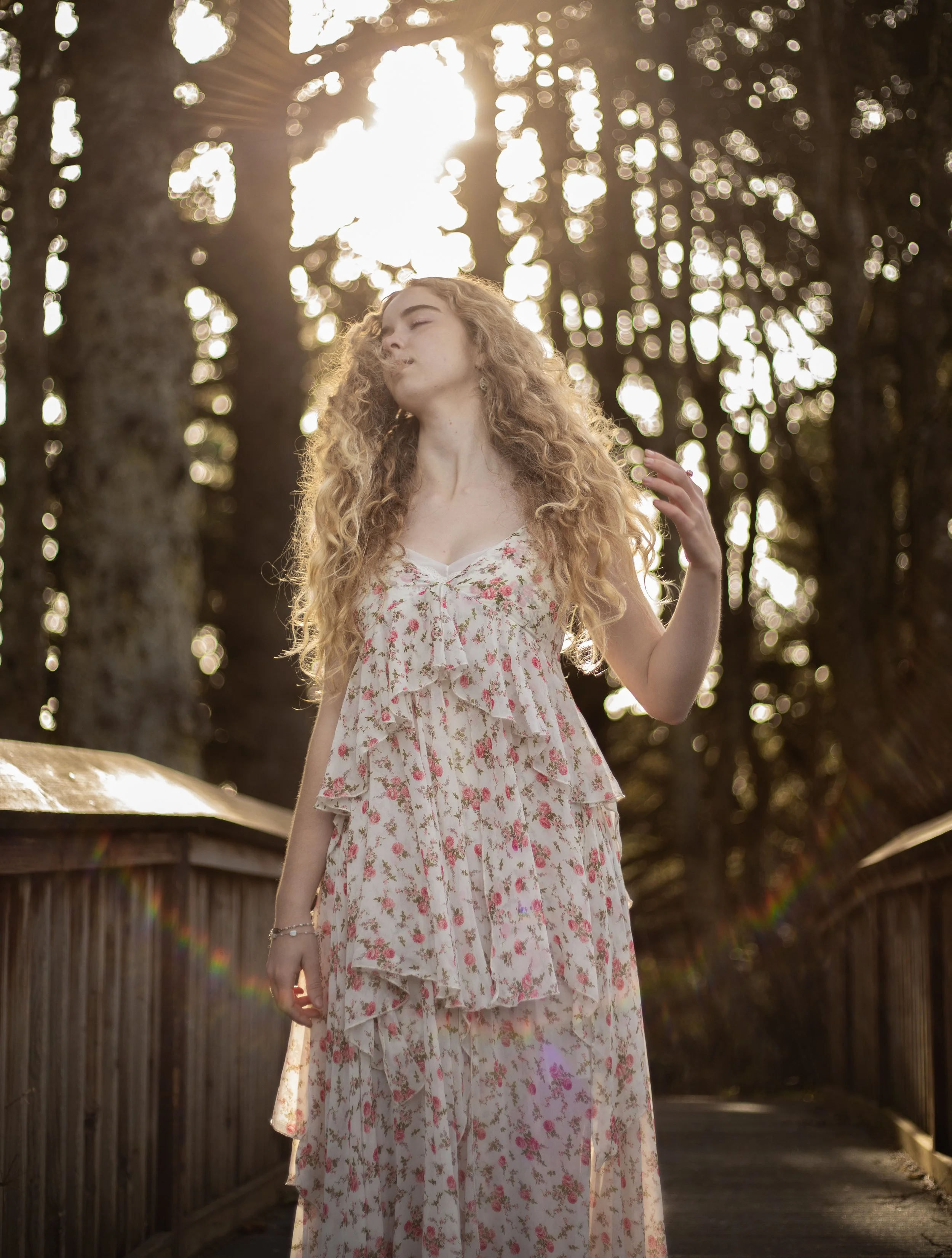 A woman with long curly blonde hair in a floral dress standing outdoors on a wooden bridge, backlit by sunlight filtering through trees.