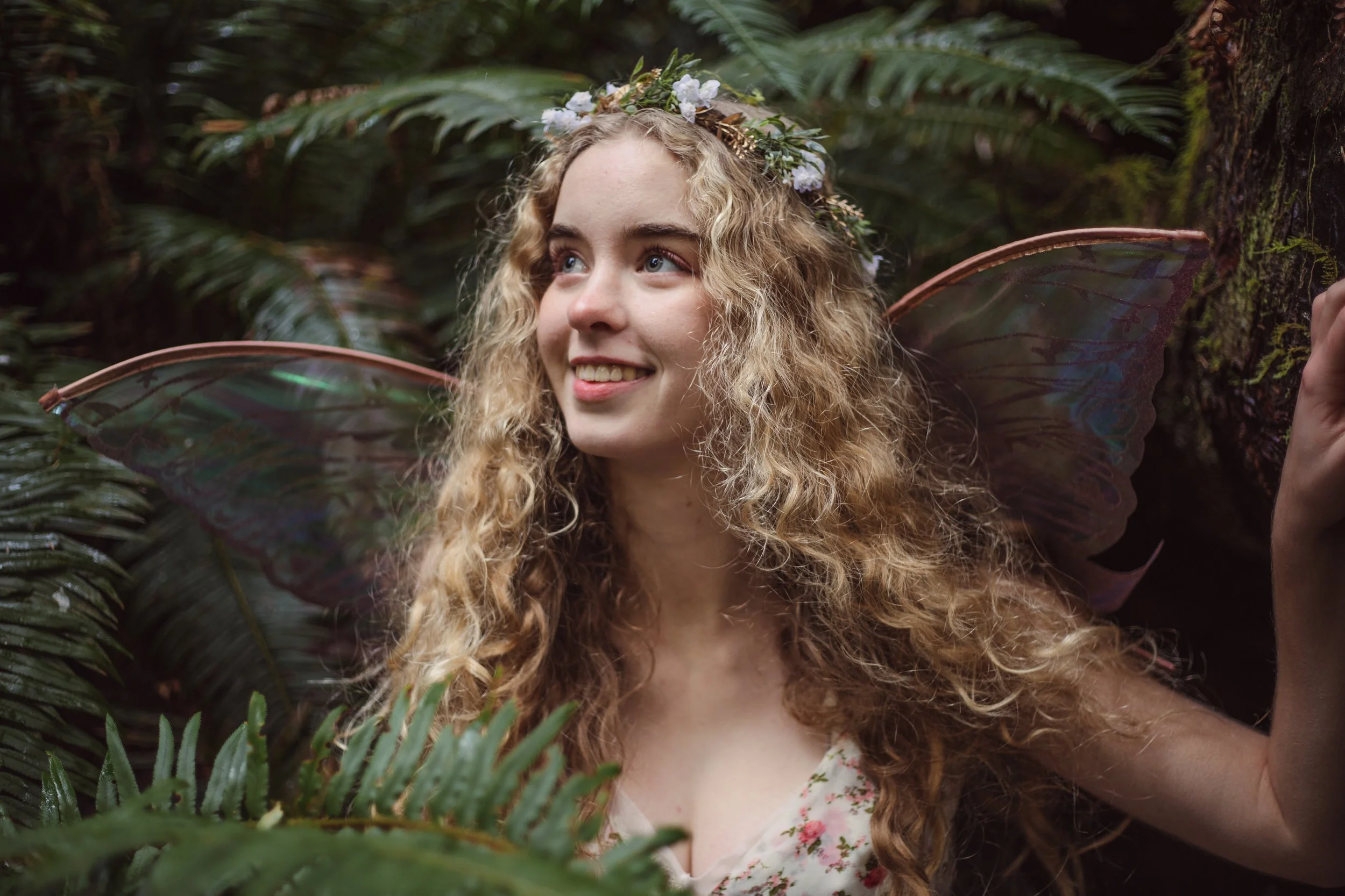 A woman dressed as a fairy or mythical creature, with curly blonde hair, floral crown, fairy wings, and a floral dress, standing in a lush green forest.