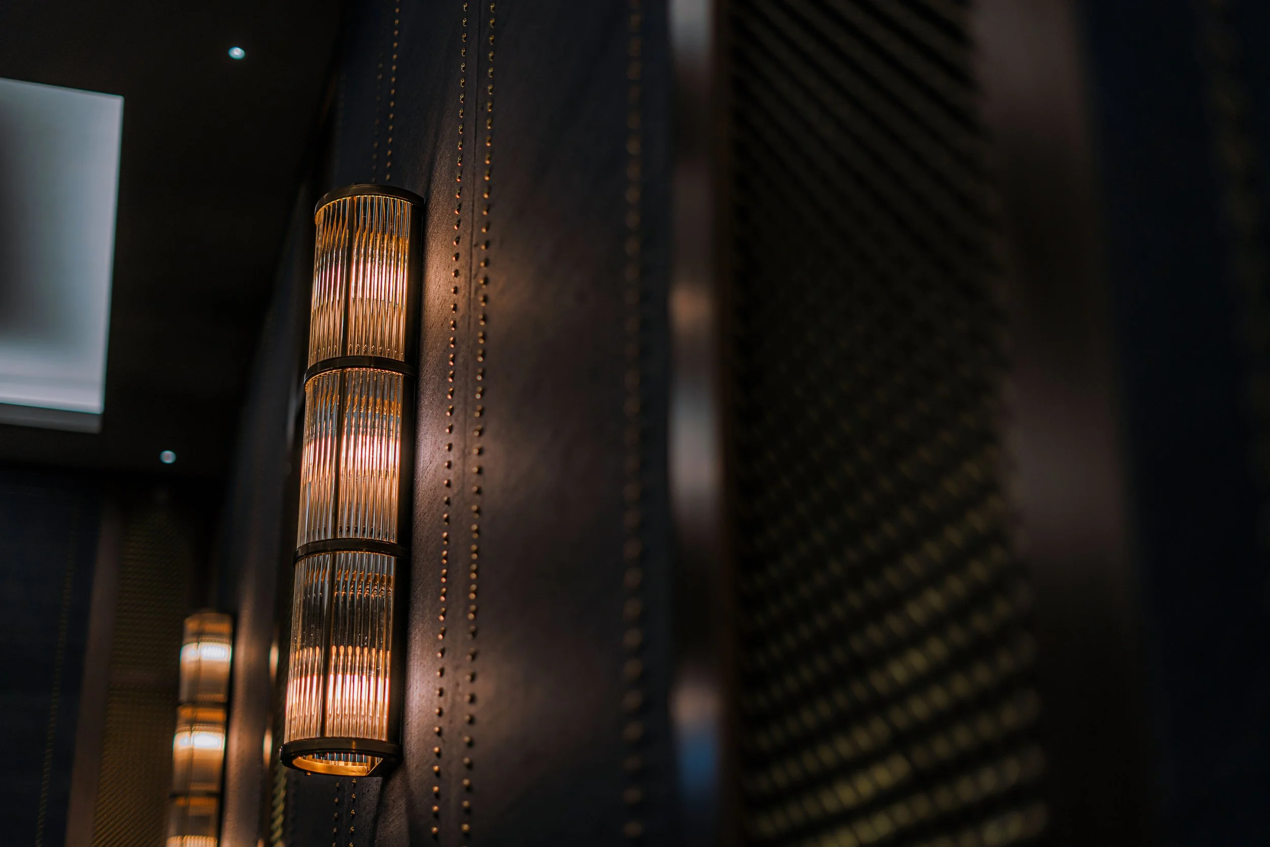 Elegant wall-mounted cylindrical lights on a dark textured wall with studded detailing, creating a modern and sophisticated ambiance.