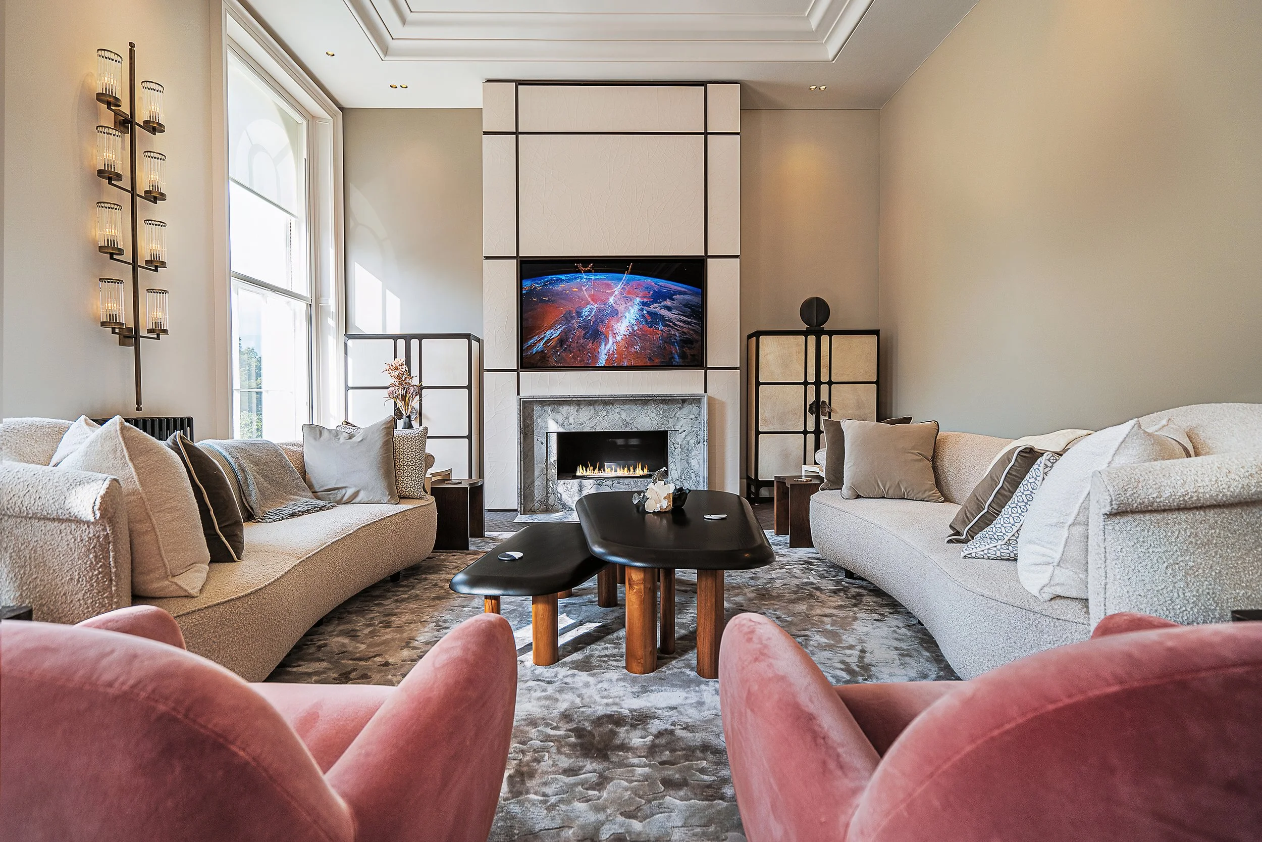 Modern living room with curved beige sofas, pink armchairs, and a central black coffee table. A fireplace with a large TV above displays an image of Earth. Soft lighting and decorative panels complete the contemporary design.