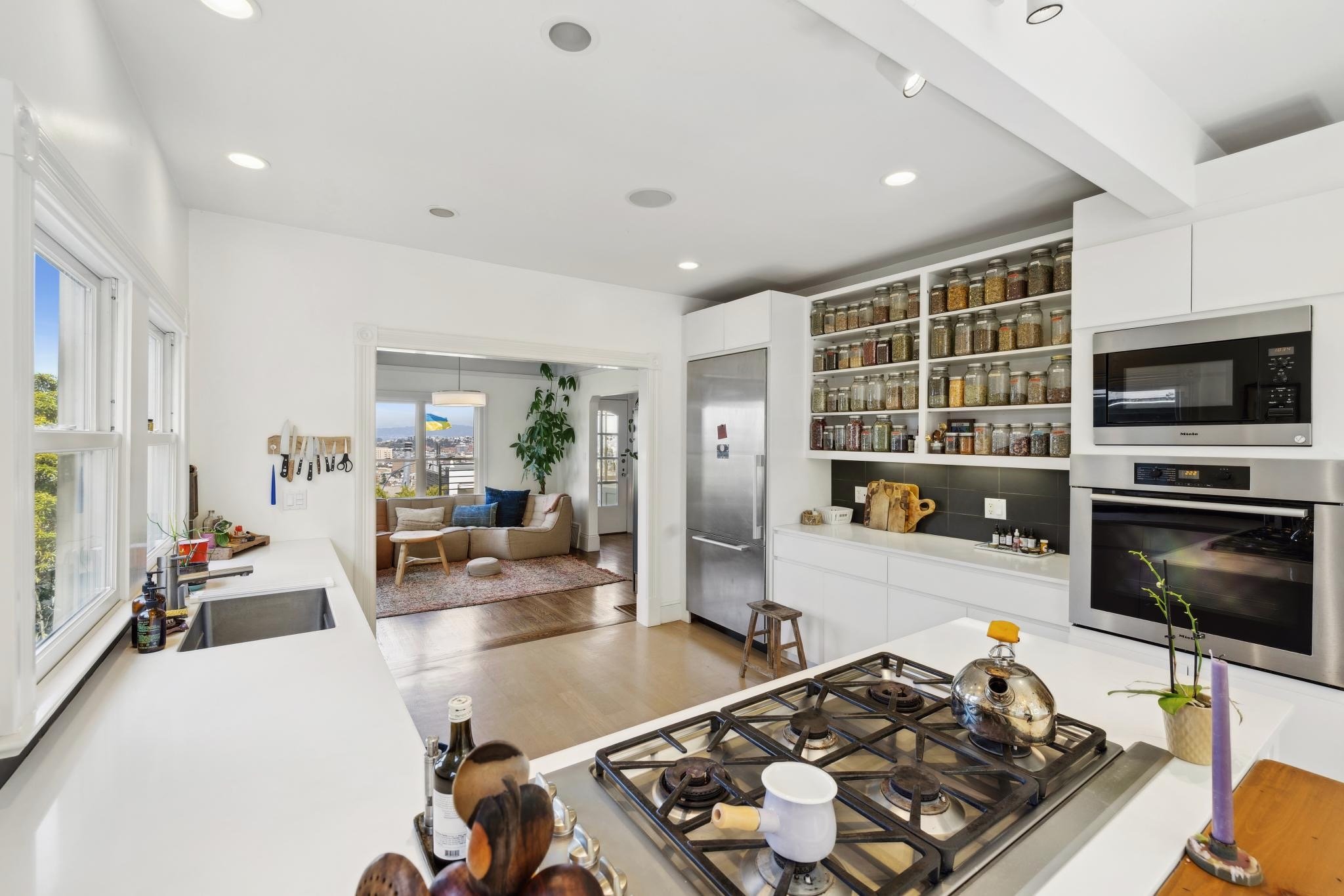 Modern kitchen with white countertops, stainless steel appliances, and open shelving filled with jars. In the background, a living room with a sofa, colorful cushions, large window, and potted plant is visible.