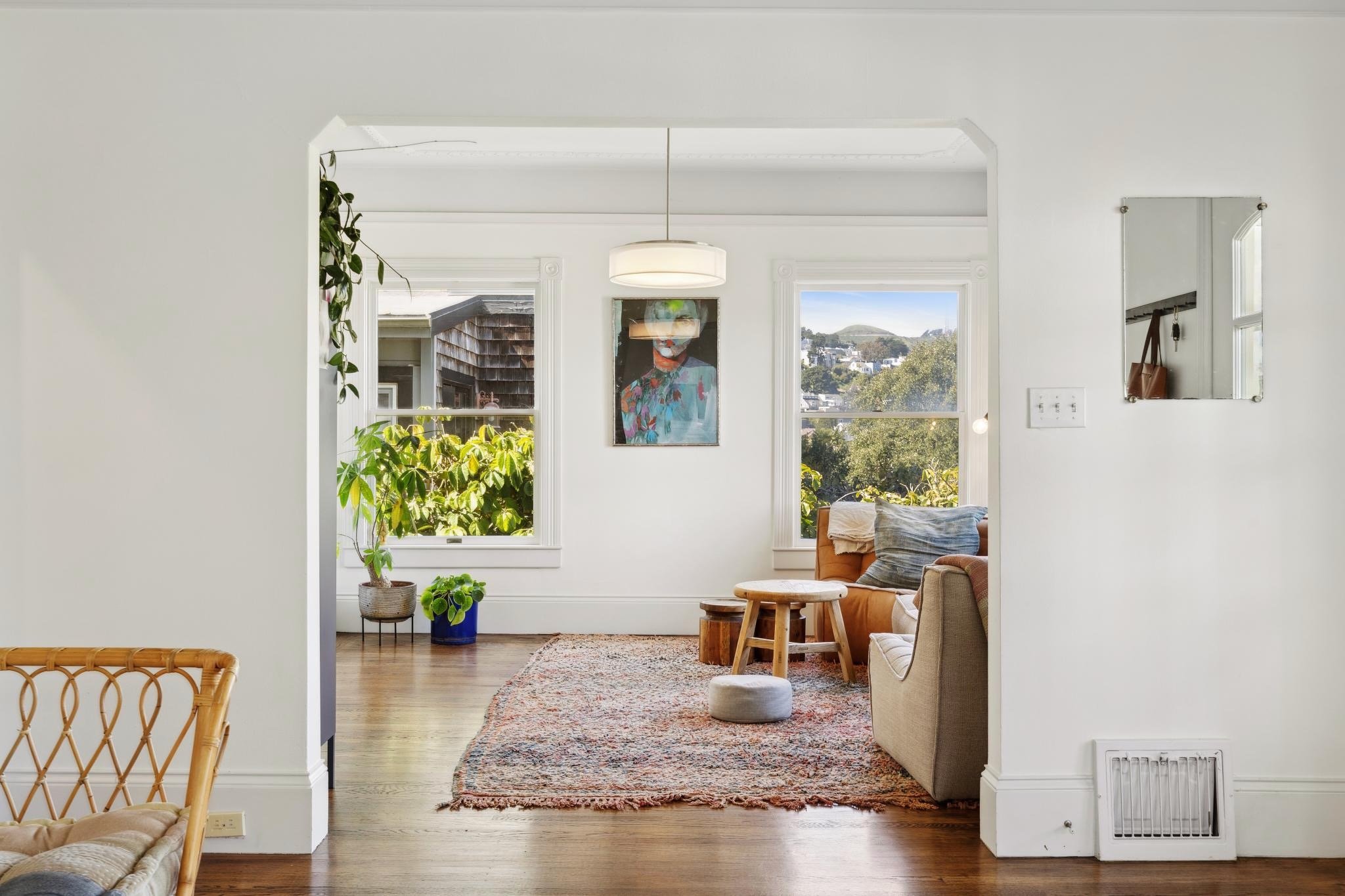 Bright living room with a large window, a cushioned couch, wooden coffee table, indoor plants, and a colorful abstract painting on the wall. There's a patterned rug on the wooden floor and a pendant light hanging from the ceiling.