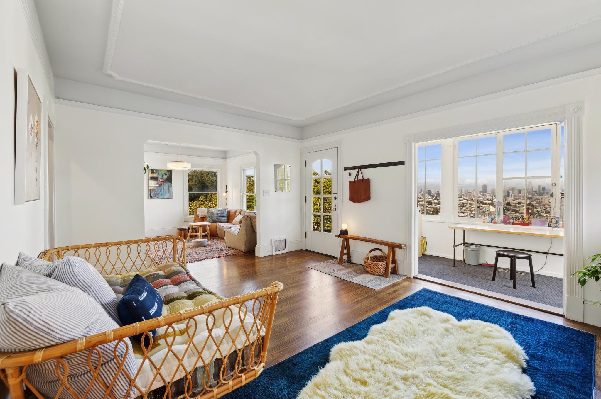 Modern living space with a wicker daybed, colorful pillows, wooden flooring, and large windows offering a city view. Includes a separate sitting area with a couch and armchairs, a cozy rug, and a door leading to a balcony. Natural light fills the roo