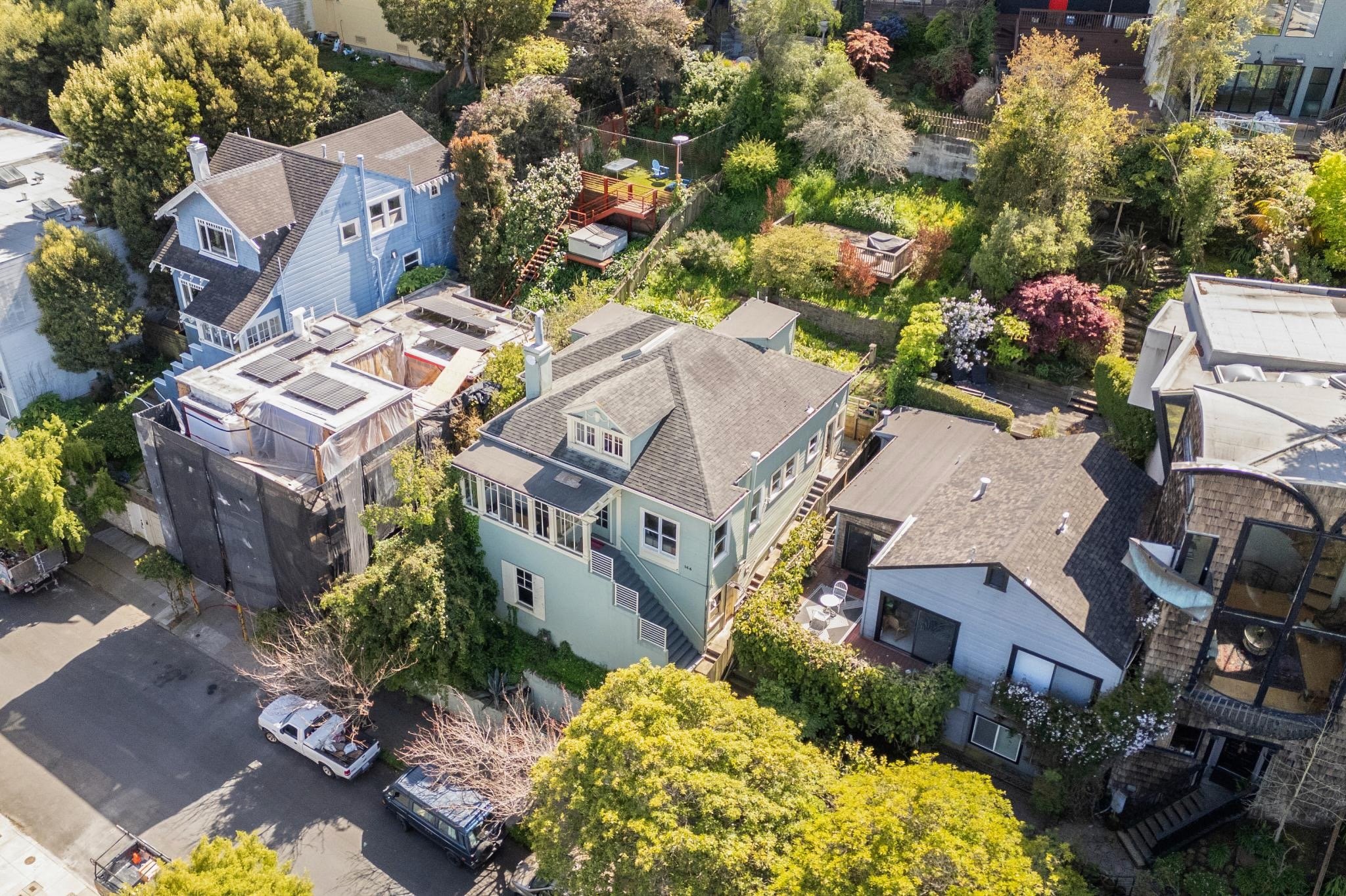 Aerial view of a residential neighborhood with multiple houses, gardens, and trees. One house is under construction with scaffolding and tarps. The scene is lush with greenery and diverse plants.
