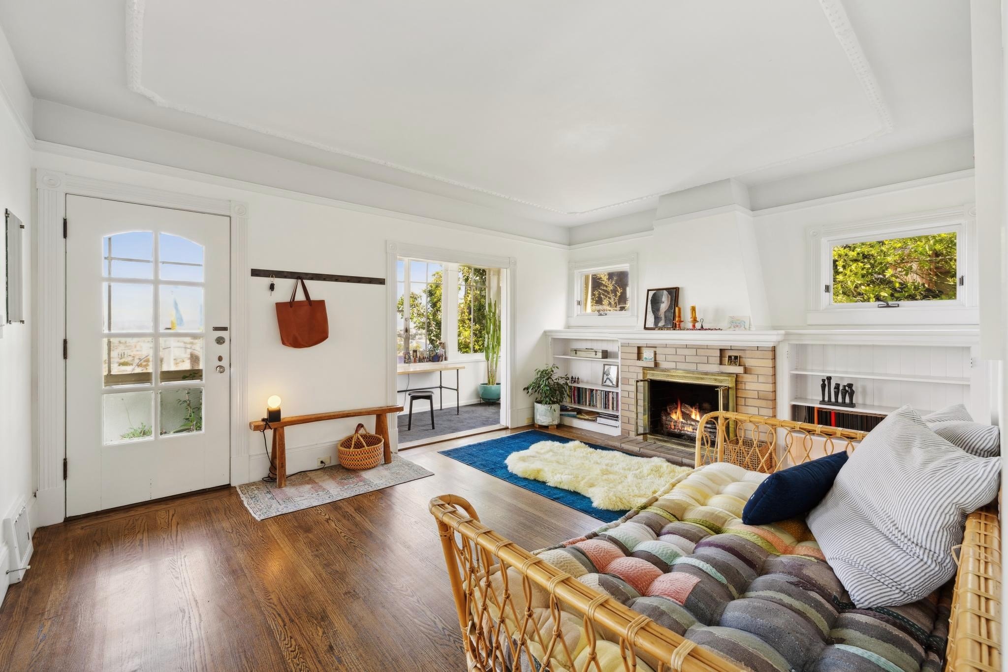 Cozy living room with a wicker couch, patterned cushions, and a fireplace. Wooden floors and white walls feature a door, bench with a basket underneath, and a window with outdoor views. Shelves filled with decor and a blue rug enhance the welcoming s
