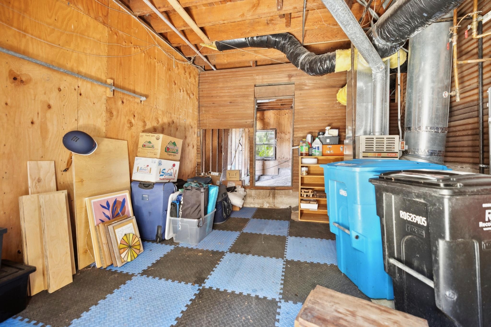 Garage interior with storage boxes, luggage, plastic bins, wooden panels, and HVAC system components. The floor is covered with blue and black foam tiles.
