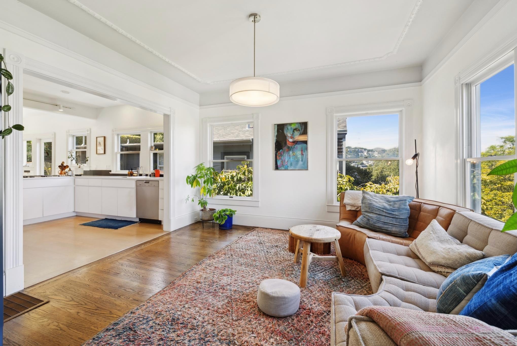 Modern living room with a brown leather sofa, colorful rug, and indoor plants, leading to a white kitchen with large windows and pendant lighting.