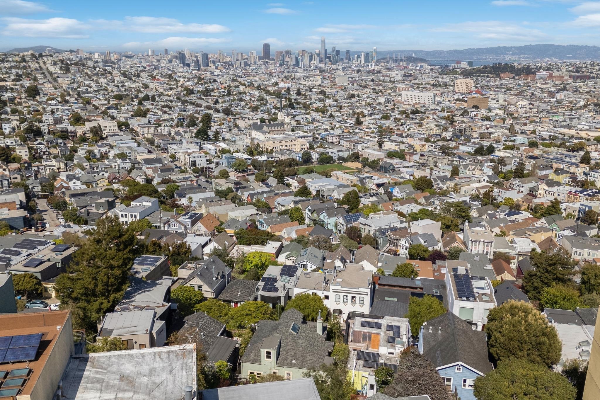 Aerial view of a densely populated urban neighborhood with rows of colorful houses and tree-lined streets, leading towards a distant skyline of a major city under a clear blue sky.