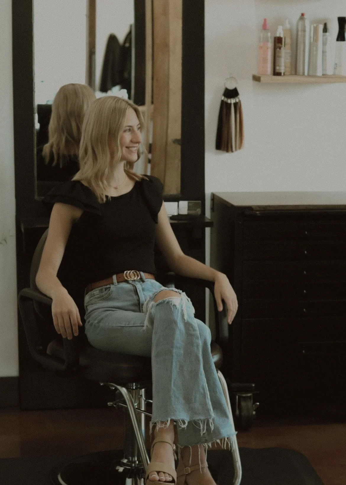 A young woman with blonde hair sitting in a black salon chair, reflected in a mirror behind her. She is smiling and wearing a black top, ripped jeans, and beige sandals. The salon has a shelf with hair products and a wall with hanging accessories.