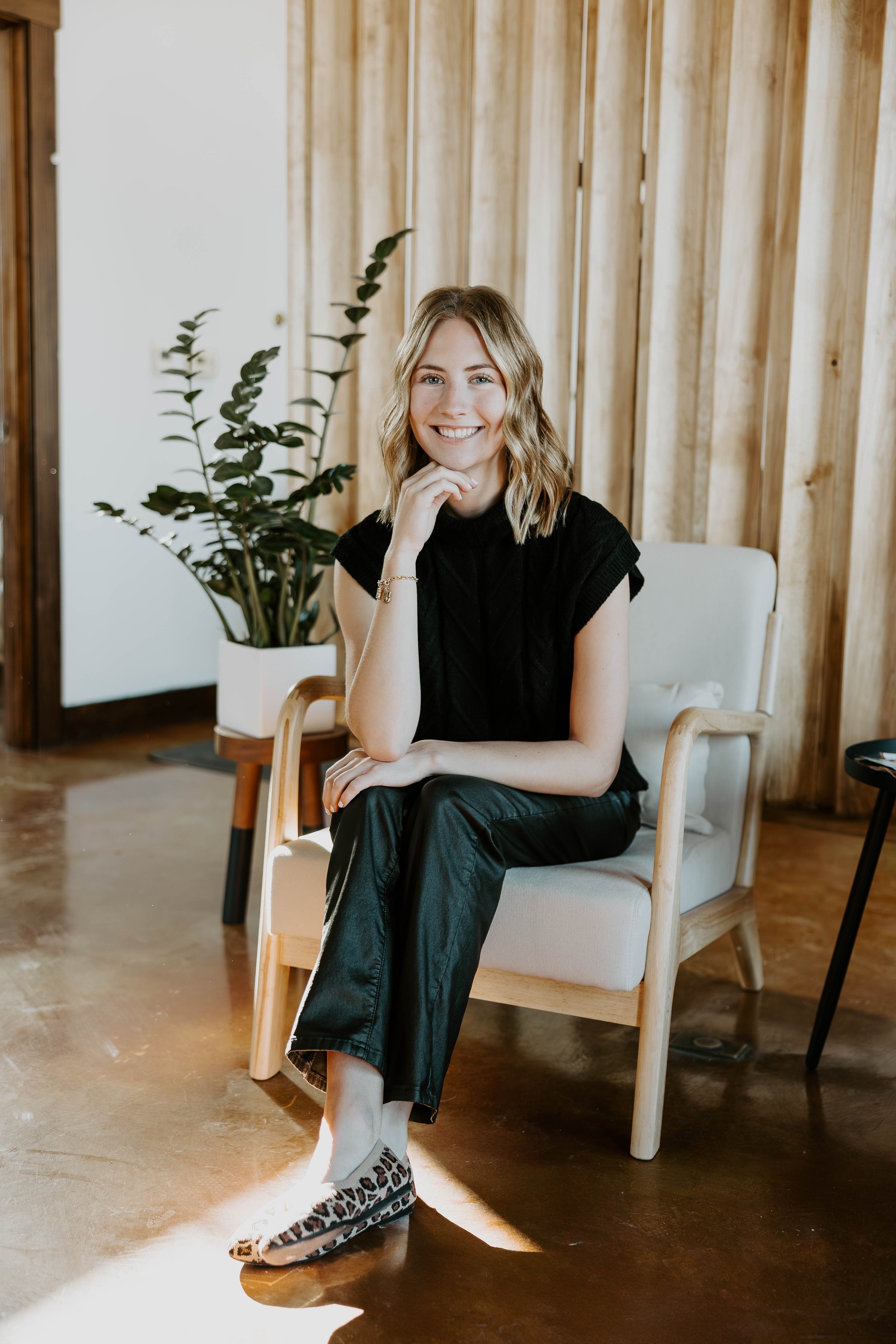 Woman in black outfit sitting in a chair indoors with a plant in the background.