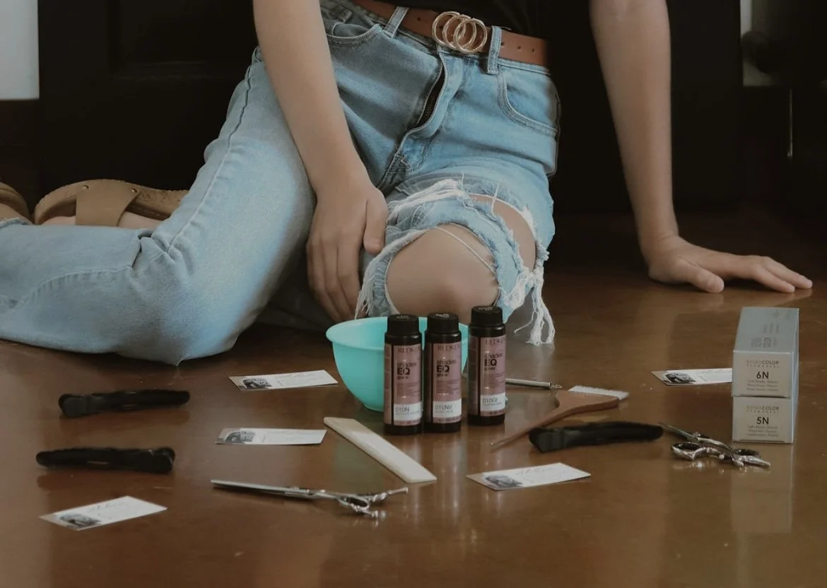 A person wearing ripped jeans and a belt is sitting on the floor surrounded by tattoo equipment including ink bottles, scissors, a spray bottle, and gloves.