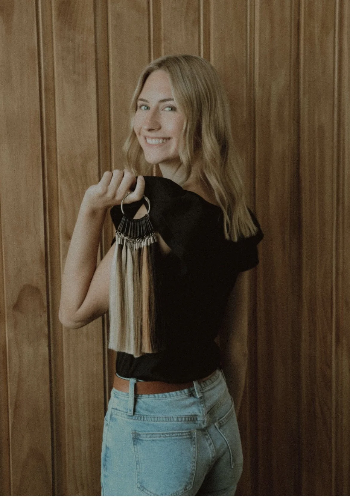 A young woman with wavy blonde hair and a big smile, holding a hair color sample fan, standing against a wood-paneled wall.