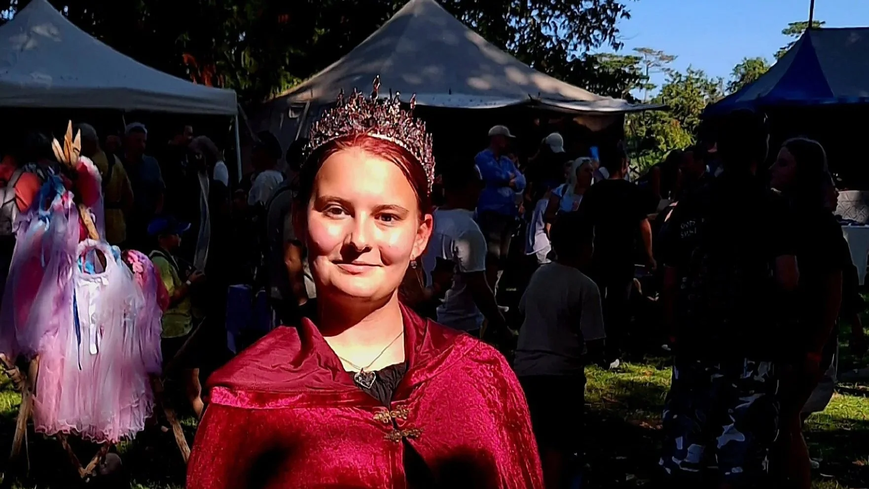Junge Frau in rotem Umhang und Krone bei einer Veranstaltung im Freien, mit mehreren Menschen und Zelten im Hintergrund.