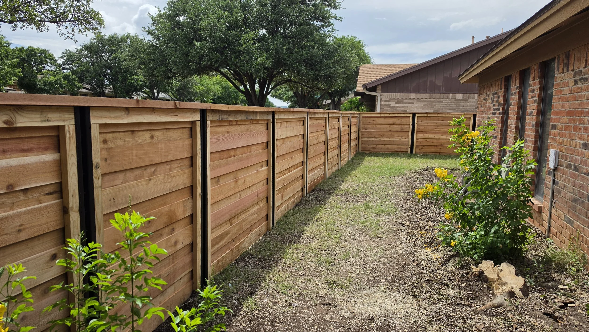 A backyard with a new wooden fence along the side of a brick house, with small yellow flowering plants and green shrubs next to the house, and large trees with green leaves in the background under a partly cloudy sky.