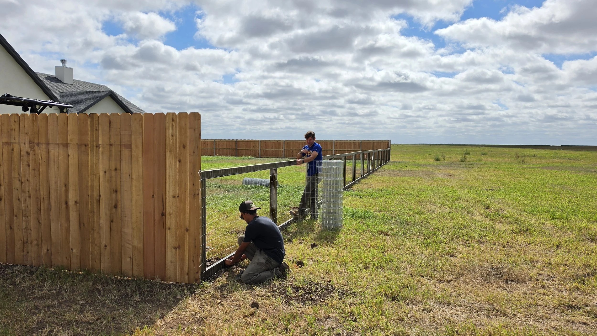 Two men building a wooden fence in an open grassy field with a house and partly cloudy sky in the background.