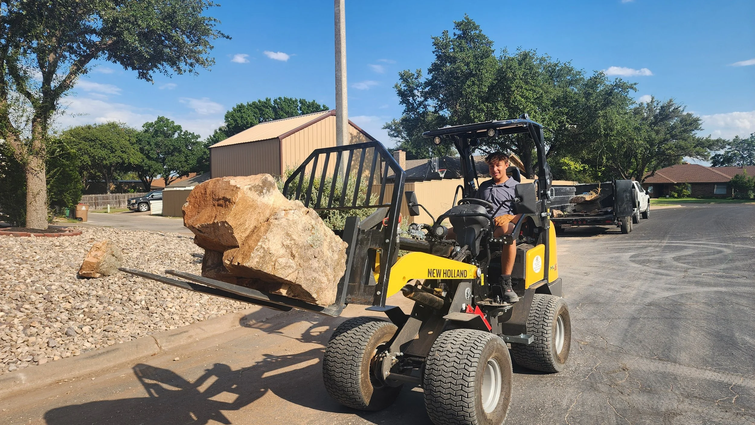A young boy sitting on a yellow New Holland compact utility tractor, holding a large rock in the front loader attachment, in a residential neighborhood with trees and houses in the background during daytime.