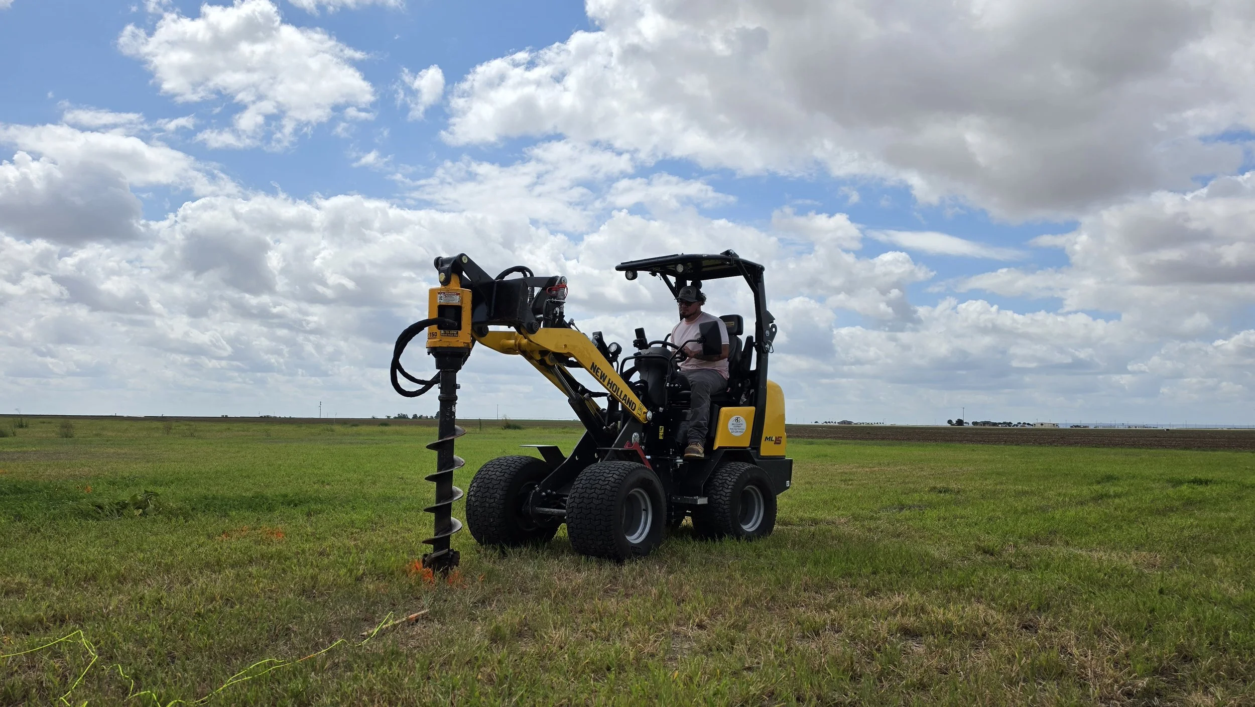 A person operating a yellow New Holland ML55 mini excavator in a grassy field under a partly cloudy sky.