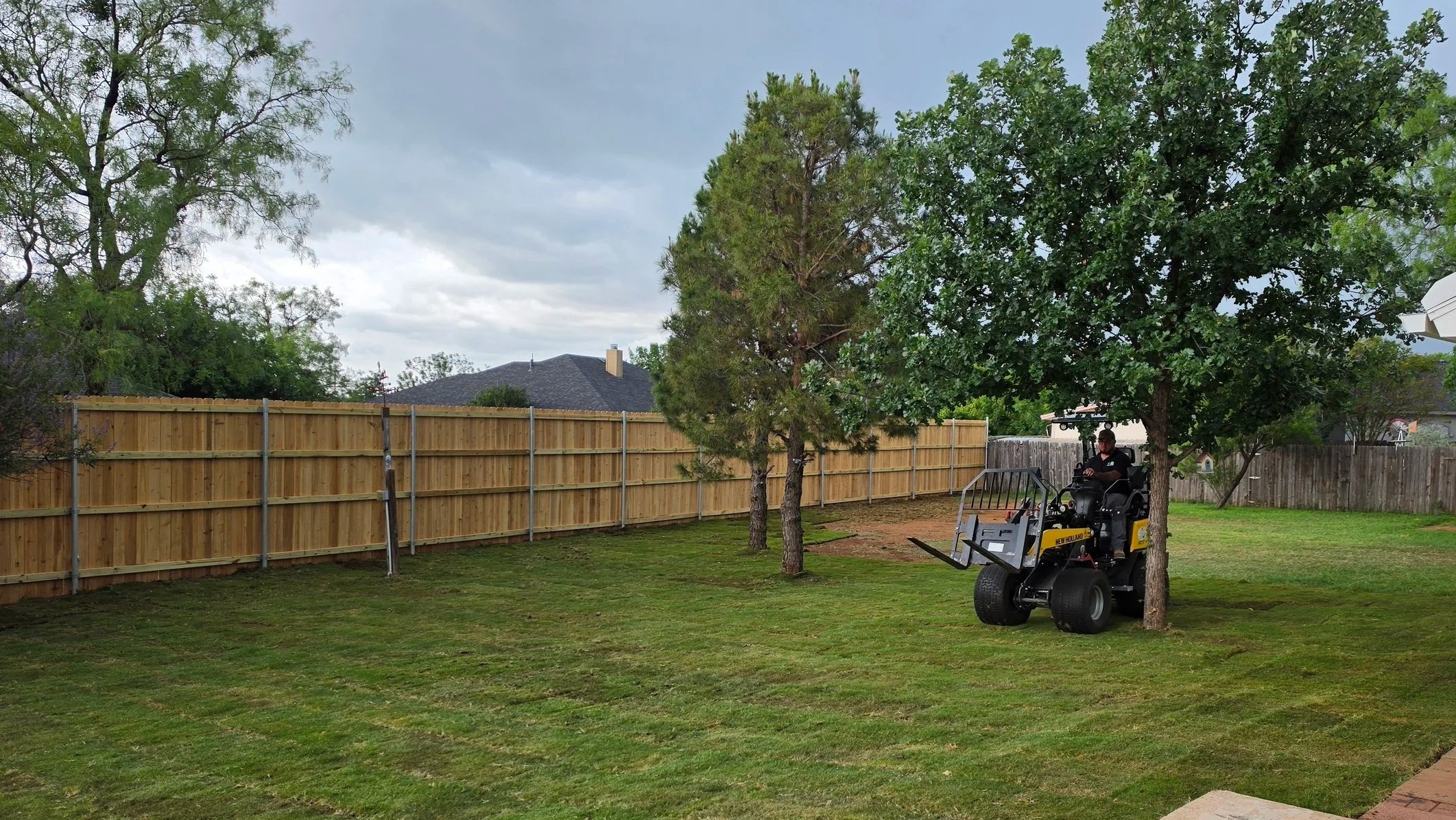 A person riding a small yellow and black lawn tractor in a backyard with a wooden fence, green grass, and multiple trees under a cloudy sky.