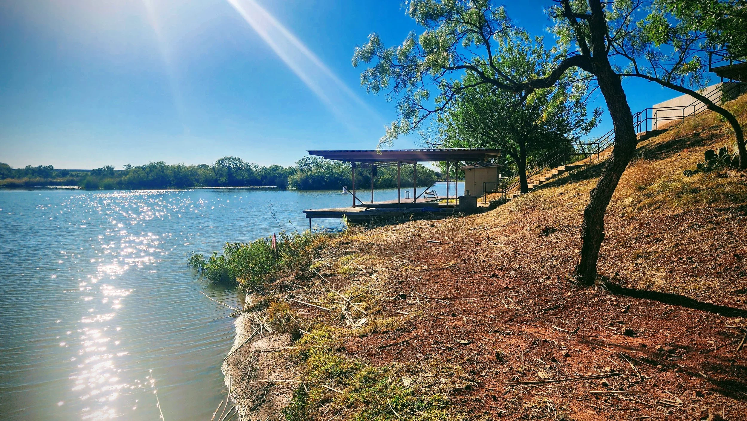 Sunshine reflecting on a calm river next to a sandy shoreline with trees and a dock with a small shelter.