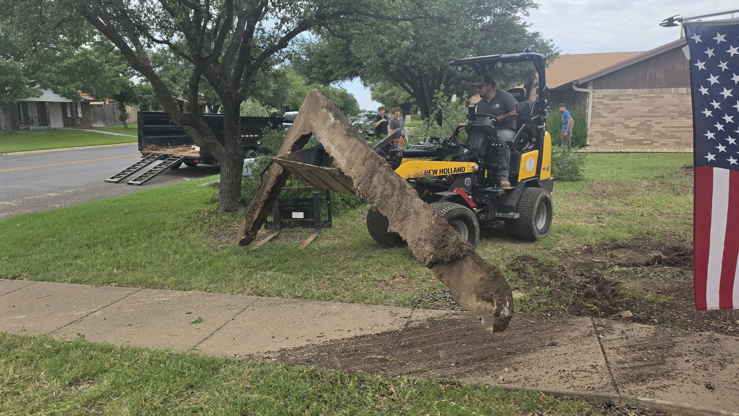 A man operating a small yellow New Holland skid-steer loader cutting down a large tree in a residential neighborhood. The tree has been partially cut and is leaning on the sidewalk, with a man and two teenagers watching nearby. An American flag is vi