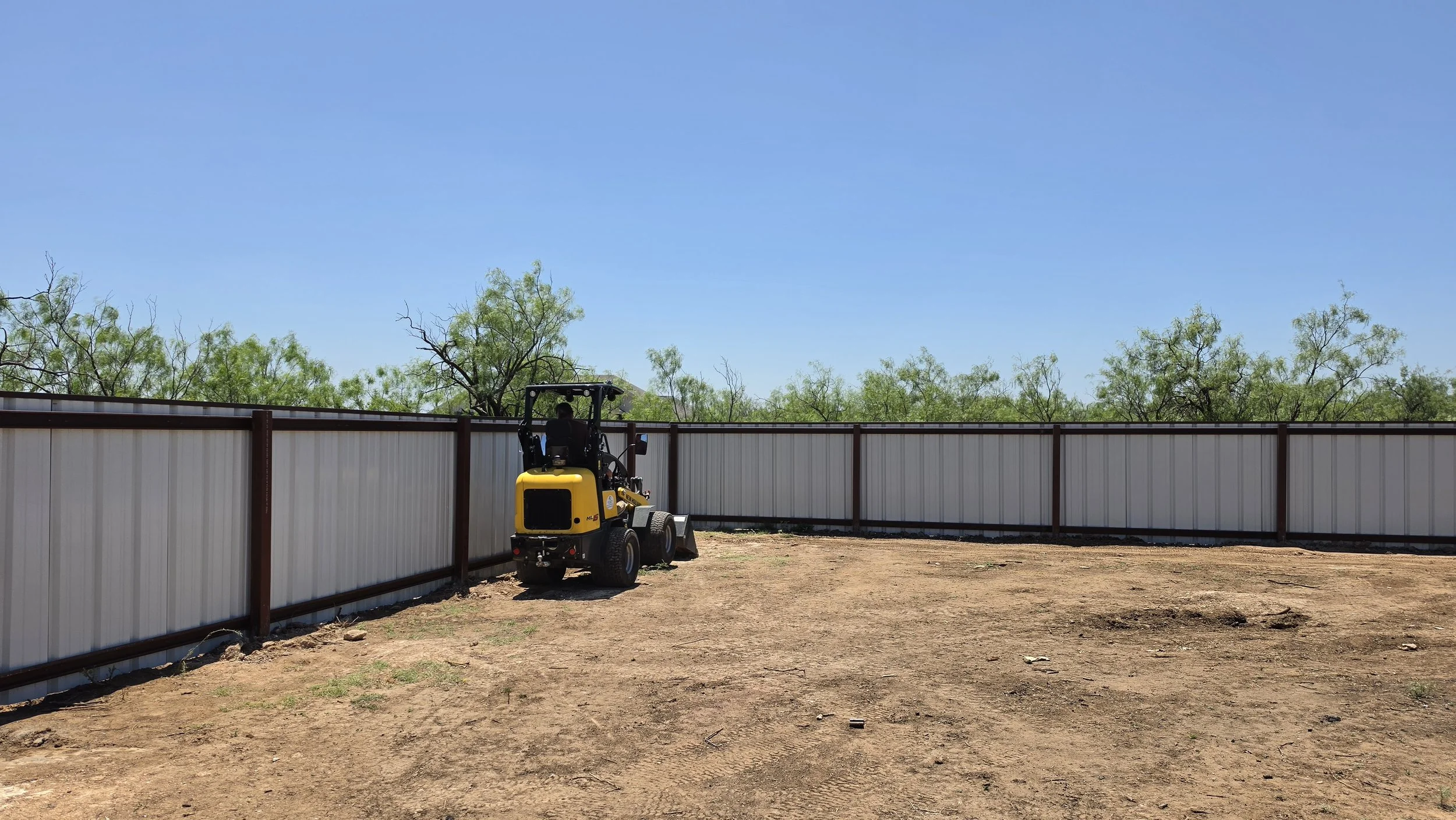 A small yellow construction vehicle parked beside a white metal fence on dirt ground with green trees and a clear blue sky in the background.