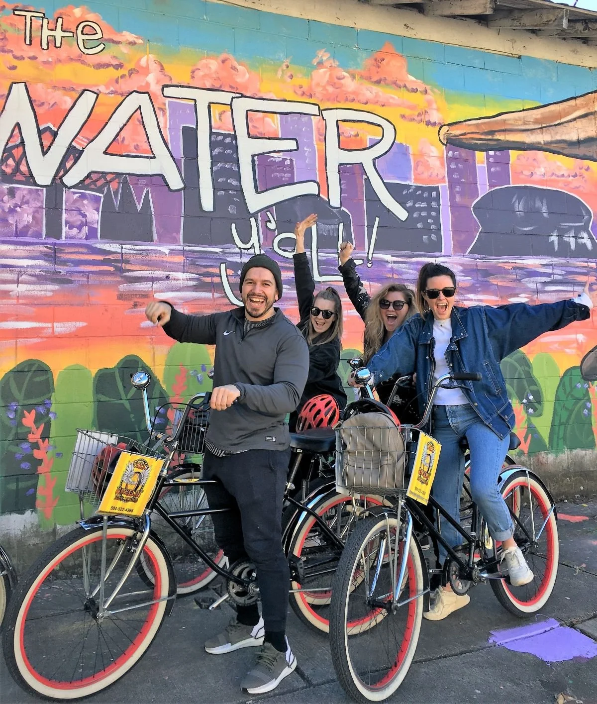 Four people smiling and celebrating with bicycles in front of a colorful mural with the words "The Water" on it.