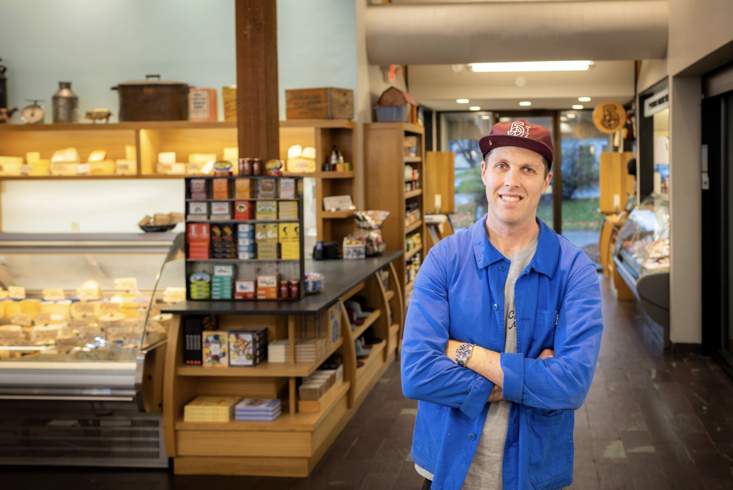 A man in a blue jacket and maroon cap stands inside a cheese shop with crossed arms, smiling, with shelves of cheese and other products behind him.