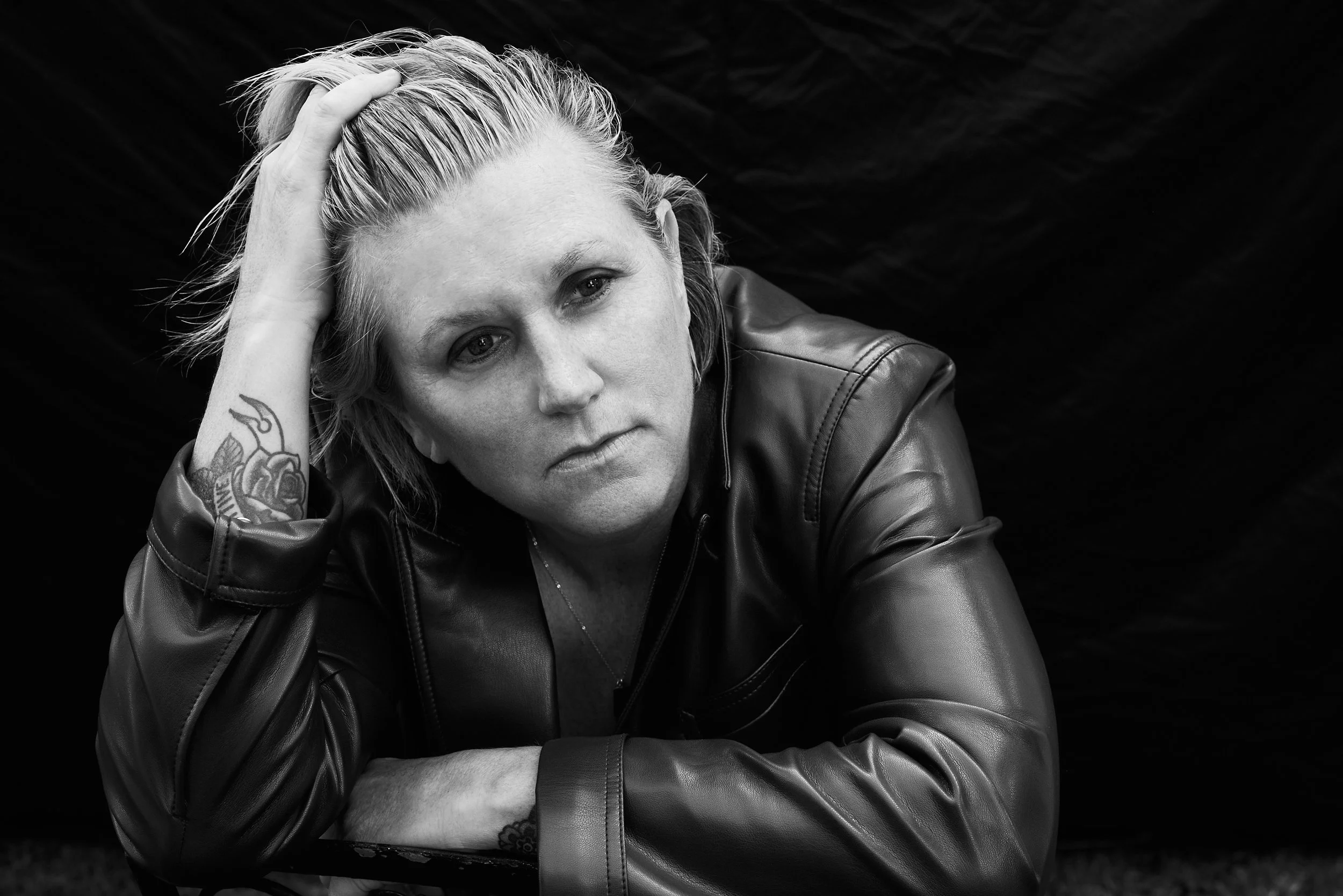 Black and white portrait of a woman with short, blonde hair, wearing a leather jacket, resting her head on her hand, with tattoos visible on her arm.