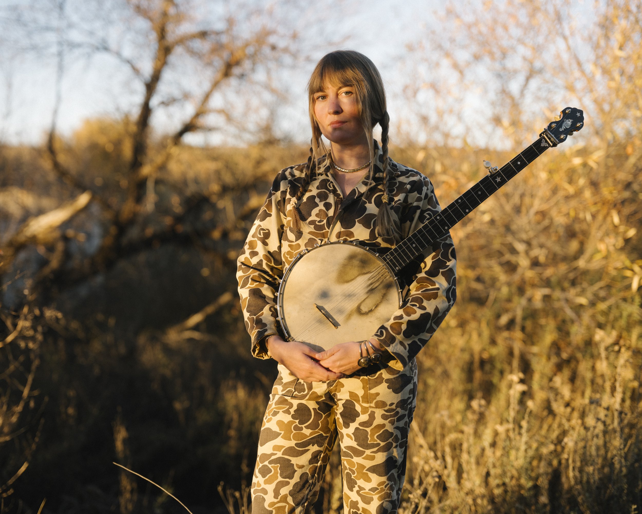 A young woman standing outdoors holding a banjo against a background of autumn trees, wearing a matching camouflage outfit