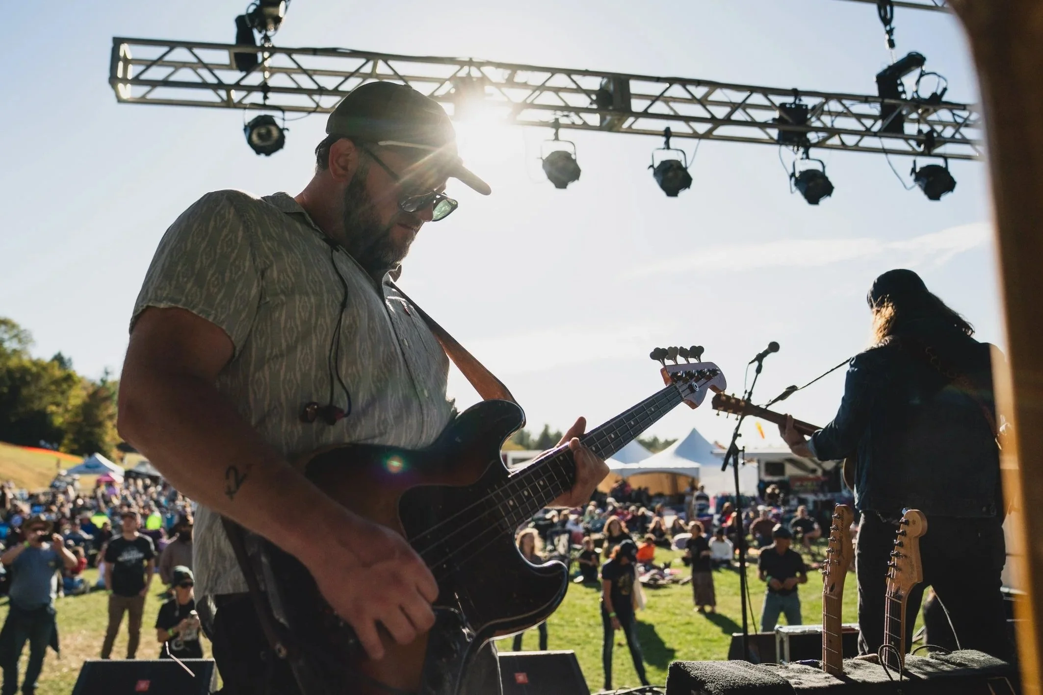 Musicians performing on stage outdoors at a festival with a crowd in the background, sunny weather, and stage lighting equipment overhead.