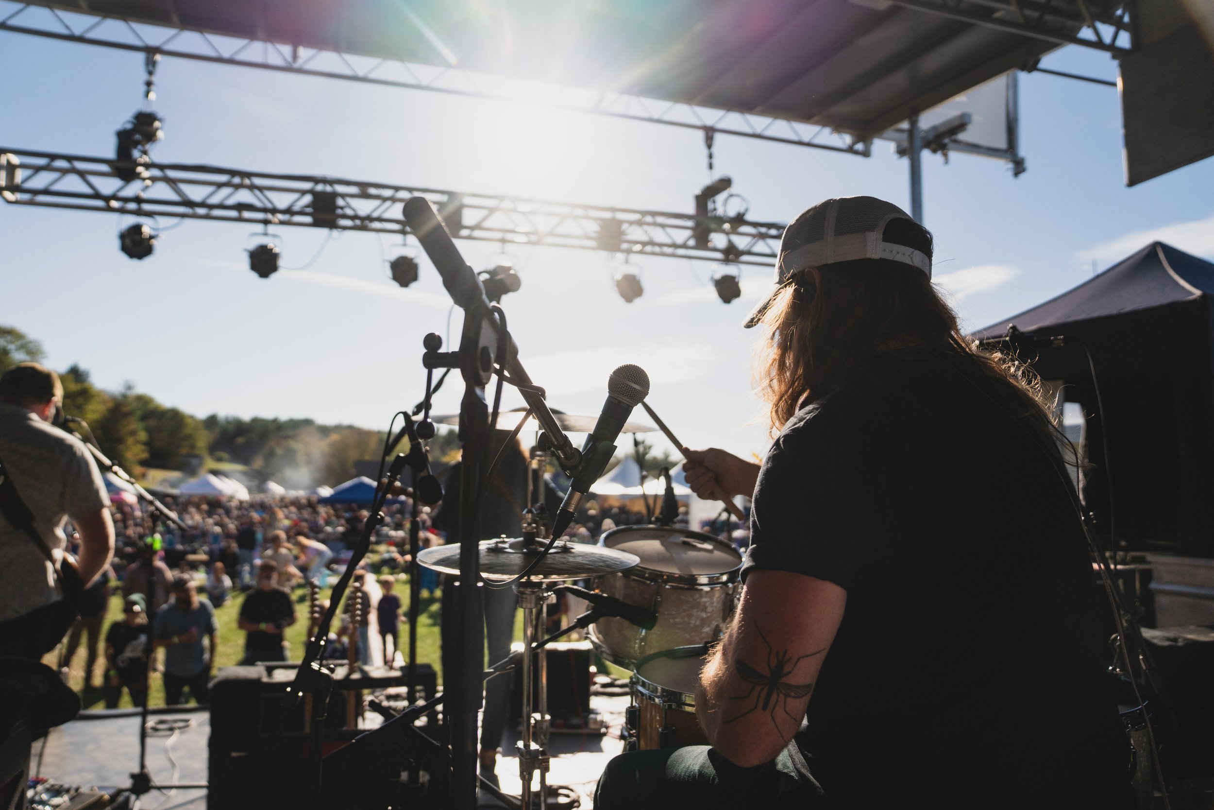 Musician on stage with drums and microphone during an outdoor concert, audience in the background, bright sunlight overhead.