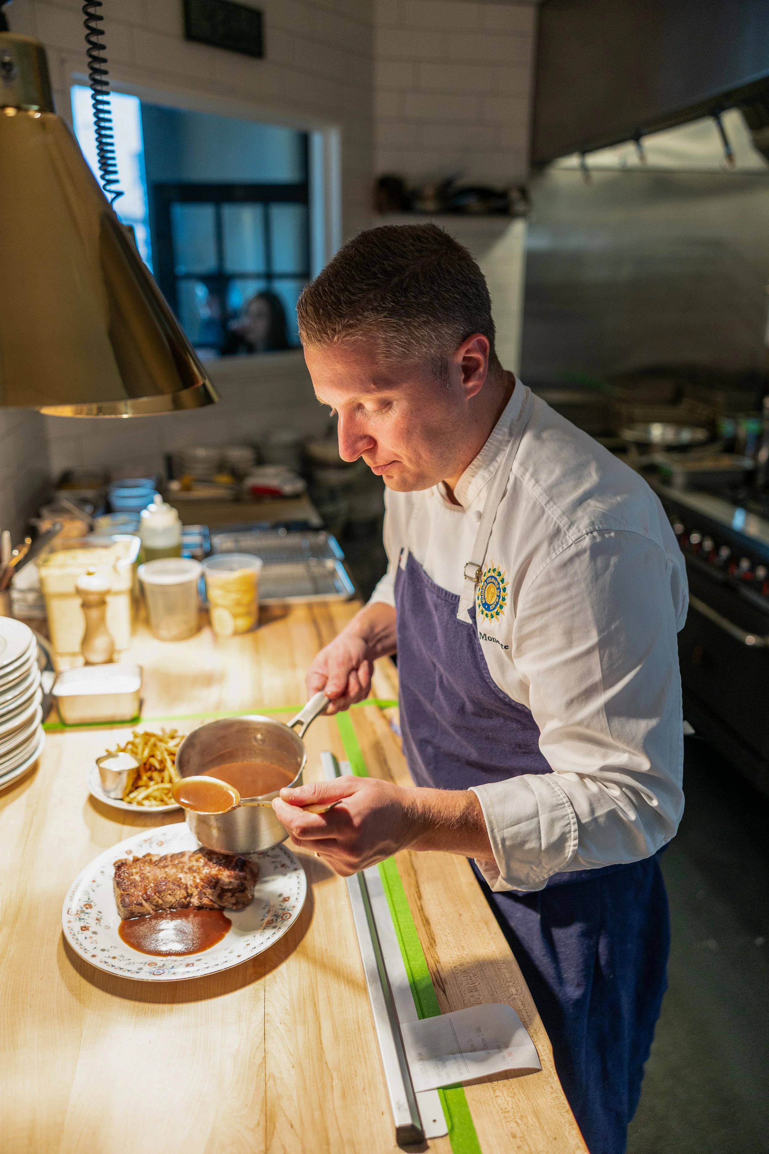 Chef pouring gravy on a cooked steak with pasta on the side in a professional kitchen.