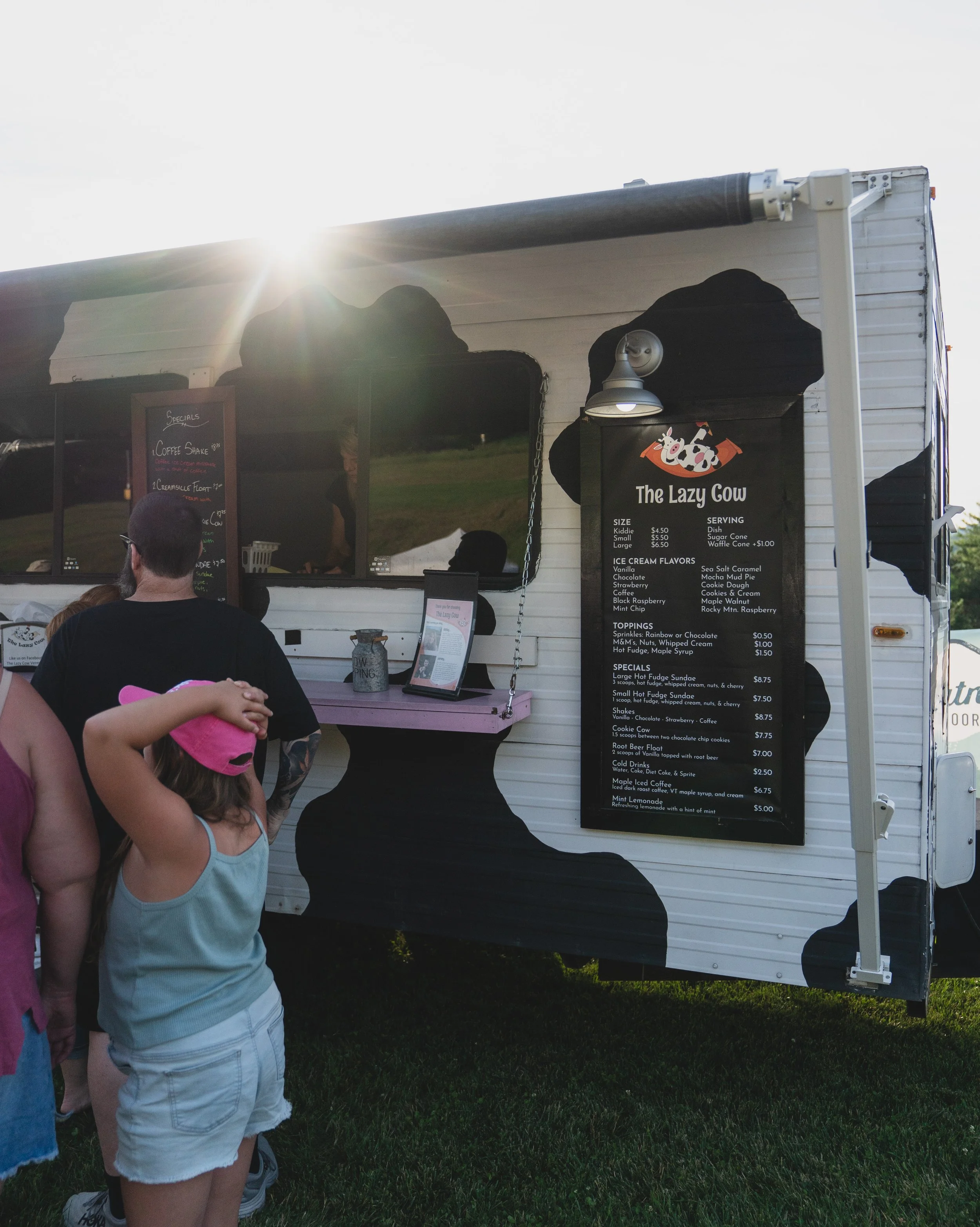 People standing in front of an ice cream truck named "The Lazy Cow" with a menu board displaying flavors and prices.