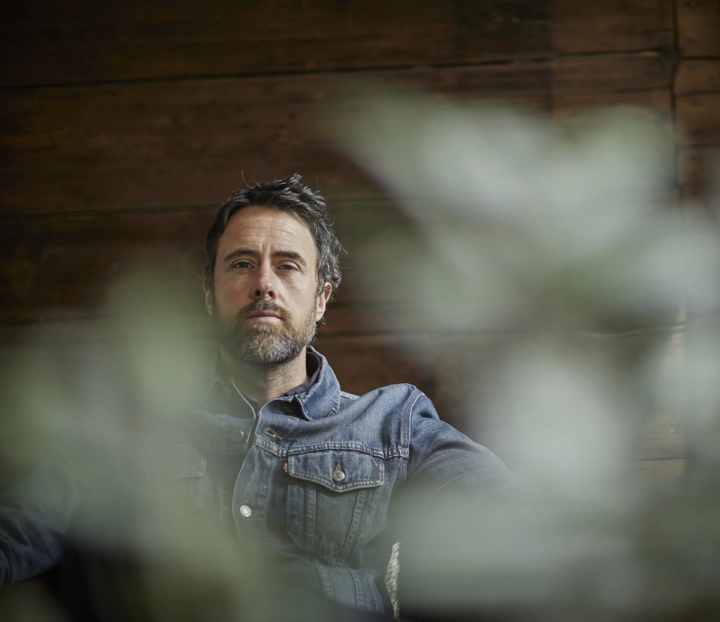 A man with dark hair and a beard, wearing a denim jacket, sitting against a wooden wall background, partially obscured by blurred green leaves in the foreground.