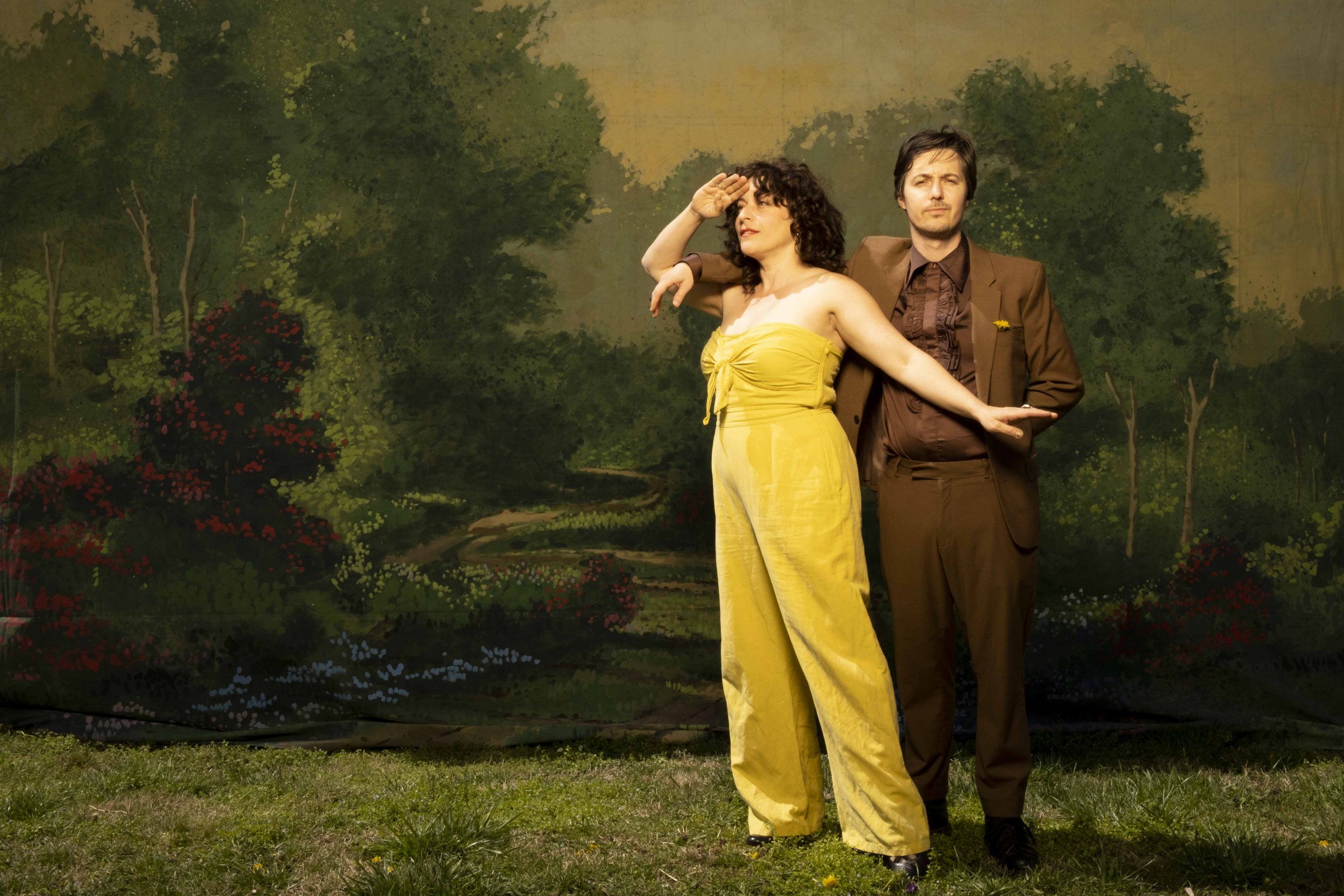 A man and woman standing on grass against a painted outdoor scene backdrop. The woman is wearing a yellow strapless jumpsuit, sculpting with one hand, and saluting with the other. The man, dressed in a brown suit, stands with one hand in his pocket and the other supporting the woman.