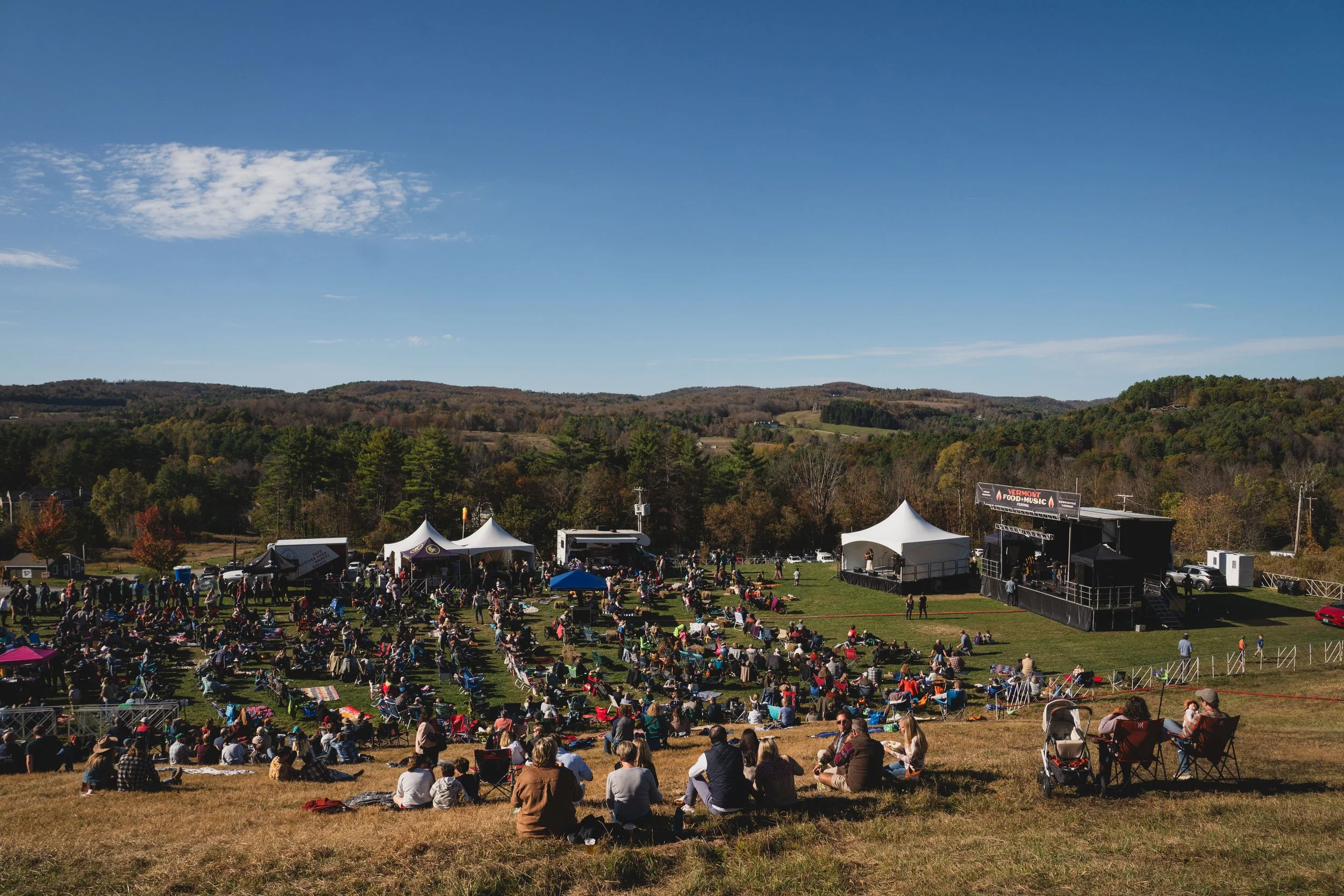 Outdoor music festival with a stage and tents, people sitting on the grass and hillside, trees, and a clear blue sky.