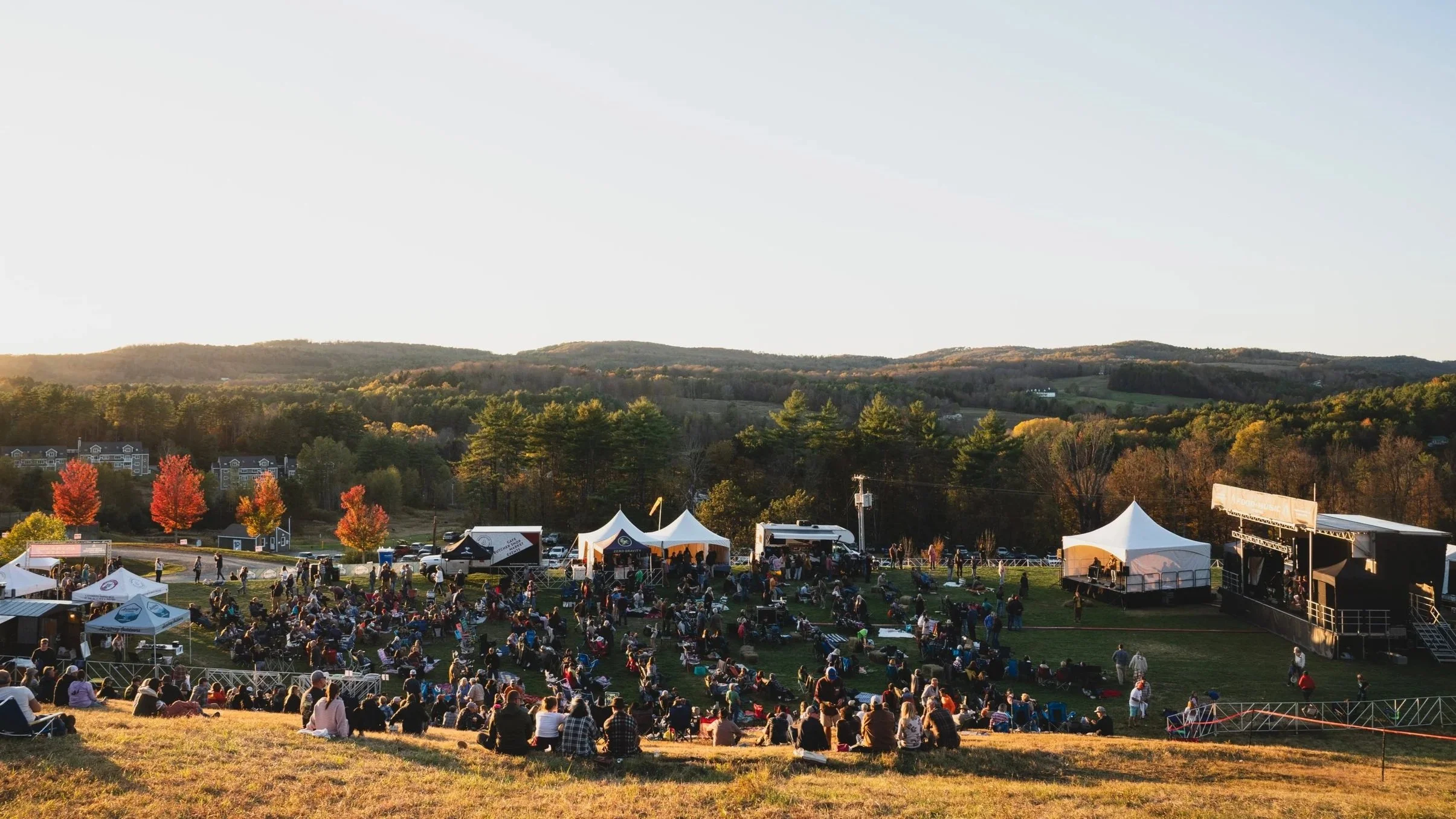 Outdoor festival scene with a stage, tents, and many people sitting and standing on a grassy hillside during sunset, with trees and hills in the background.