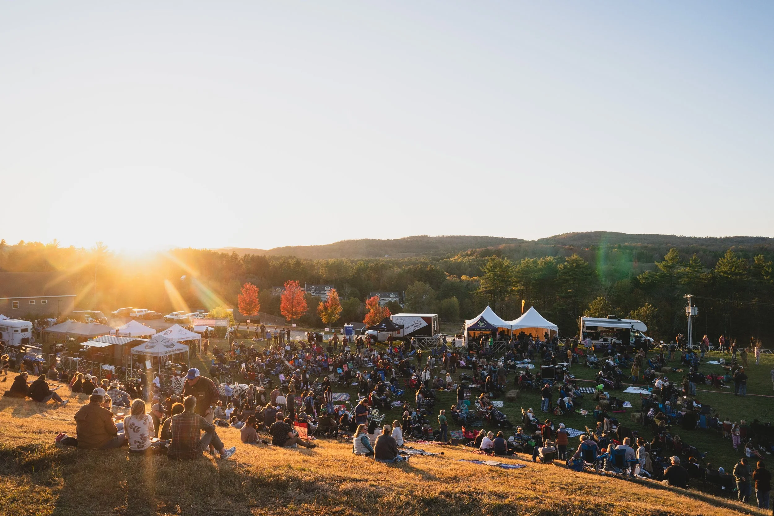 A large outdoor gathering or concert at sunset, with many people seated on the grass, some under tents, surrounding a stage area with trucks and tents in a scenic, hilly landscape with autumn-colored trees.