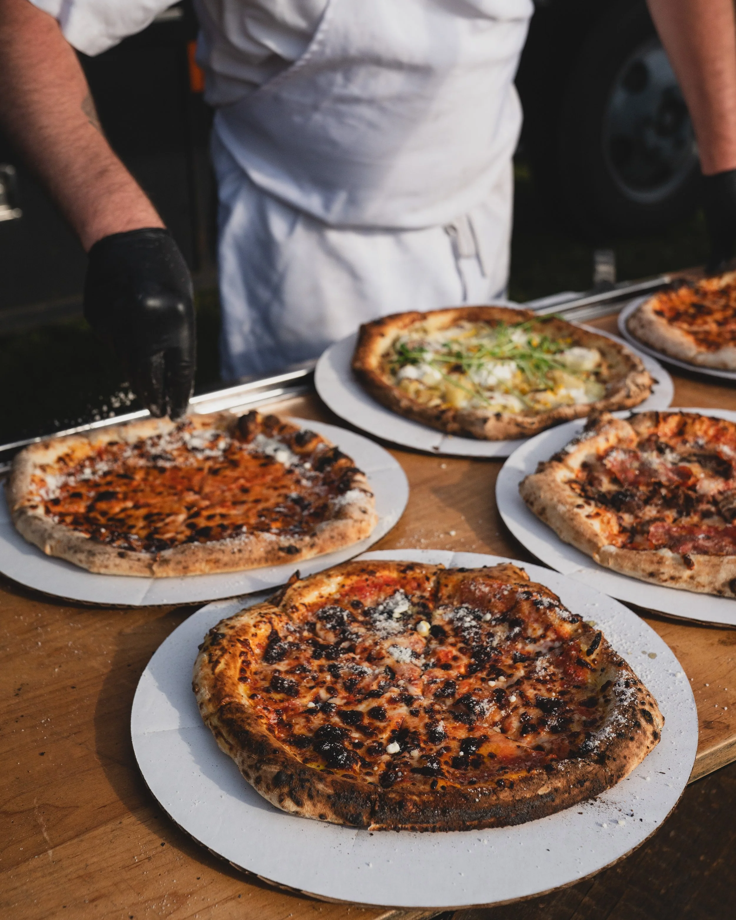 Several freshly baked pizzas on white paper plates on a wooden table, with one being sliced by a person wearing a black glove and white apron.