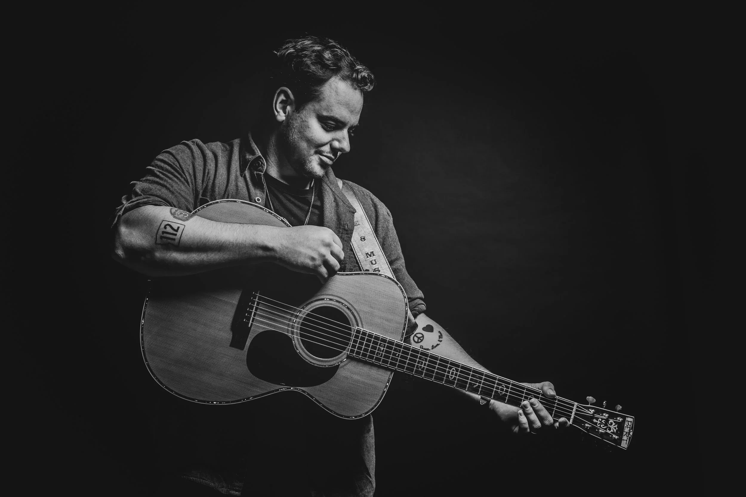 A young man playing an acoustic guitar, with a focused and content expression, against a plain black background.